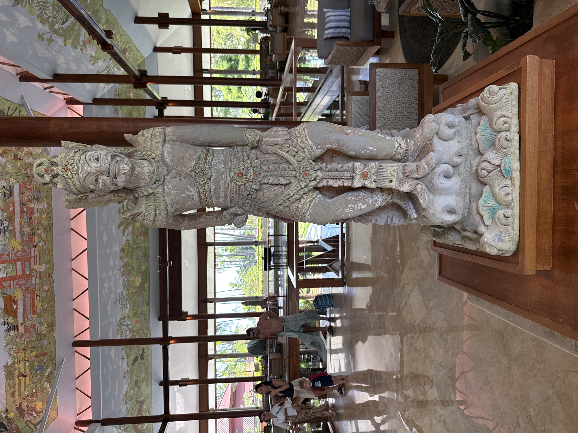 The image captures a vibrant interior space, likely a resort lobby or open-air restaurant, in Kuta Selatan, Indonesia. In the foreground, a large, weathered stone statue stands prominently on a wooden pedestal. The statue depicts a deity or mythical figure, intricately carved with traditional attire, elaborate headwear, and jewelry. Faded colors of white, red, and subtle blue/green accents are visible on its surface.
The background reveals a spacious area with high ceilings supported by substantial wooden beams. The ceiling and upper walls are adorned with extensive murals depicting lush landscapes, traditional buildings, various animals (including deer and cats), and possibly mythological narratives. These murals are subtly illuminated by soft, reddish uplighting. Large windows or open architectural sections offer expansive views of a verdant, tropical exterior, featuring numerous palm trees and distant structures, indicating a sunny daytime setting.
The floor is highly polished and reflective, mirroring the interior architecture and the people present. Several individuals are visible: a shirtless man wrapped in what appears to be a sarong or towel, a woman, and a young child wearing a red and blue shirt with some indistinct white text. Other elements include sofas with cushions and woven chairs, contributing to a comfortable and exotic ambiance. The scene suggests a blend of traditional culture, natural beauty, and modern hospitality.