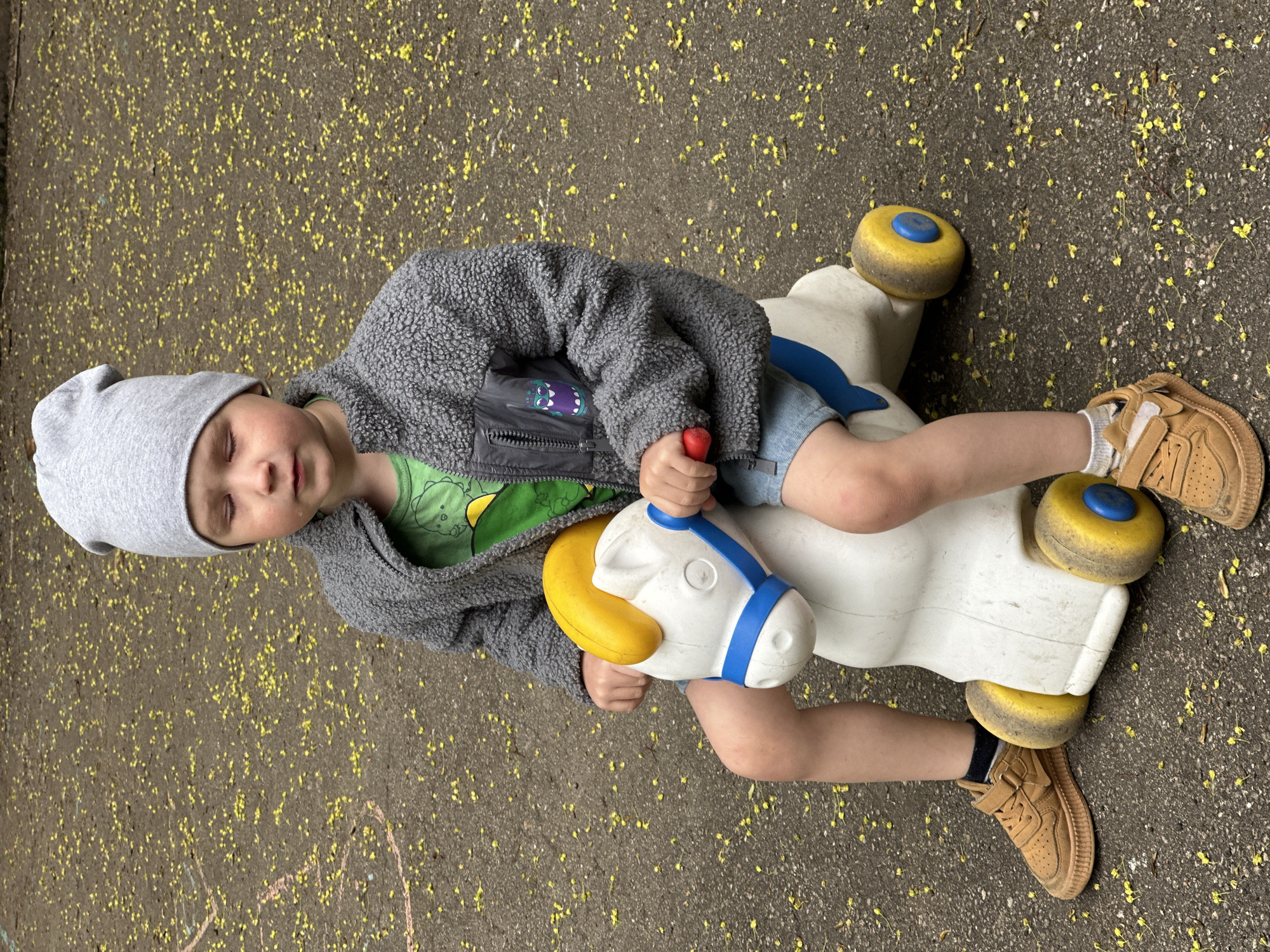 A young child wearing a gray hat, gray fleece jacket over a green T-shirt, and jean shorts is lying on a white plastic toy horse with yellow and blue wheels. The child's eyes are closed. The child is wearing tan shoes. The ground is pavement with scattered yellow flowers. The image does not contain sufficient information to determine the location in Nizhny Novgorod, Russia.
