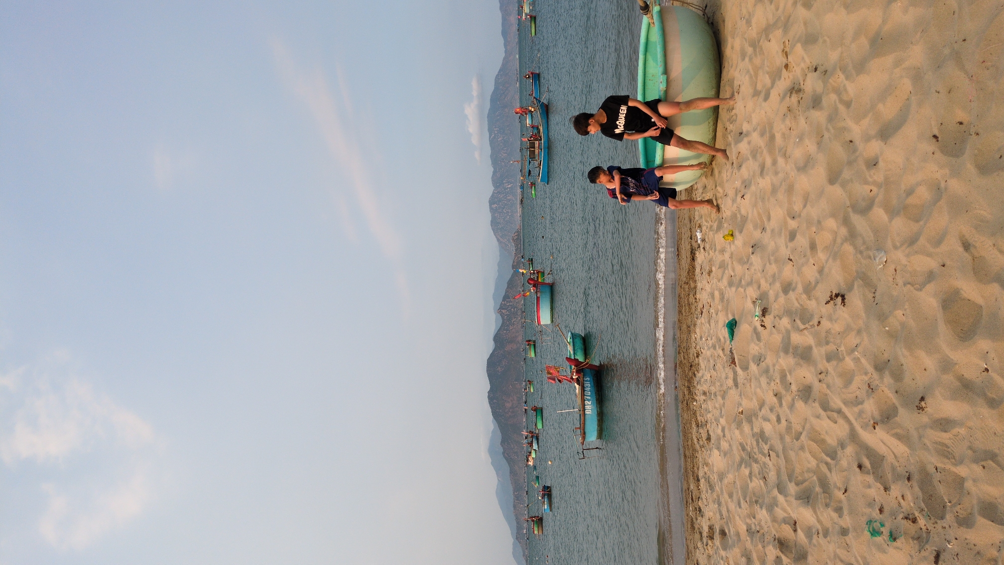 Two people are sitting on a boat. There are several boats in the water. The water is located near a beach. The image was taken in Phan Rang – Tháp Chàm, Vietnam. Mountains are in the background.