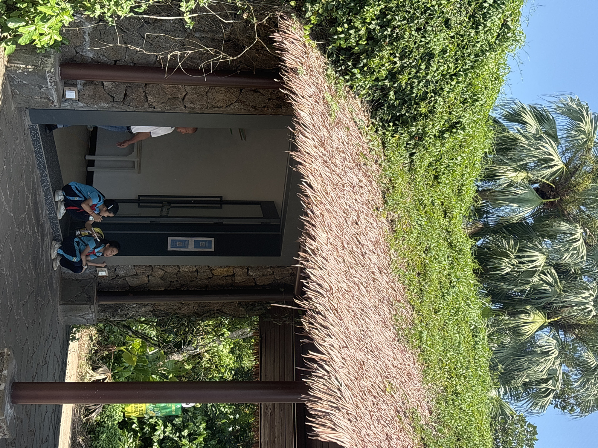 Two young girls, one wearing a blue and yellow uniform with a backpack, are crouched on the ground in front of a building with a thatched roof and stone walls. A man in a white shirt and jeans stands in the doorway of the building, looking to the left. Palm trees and lush green vegetation are visible in the background under a clear blue sky. This scene is likely located in Xiuying District, China.