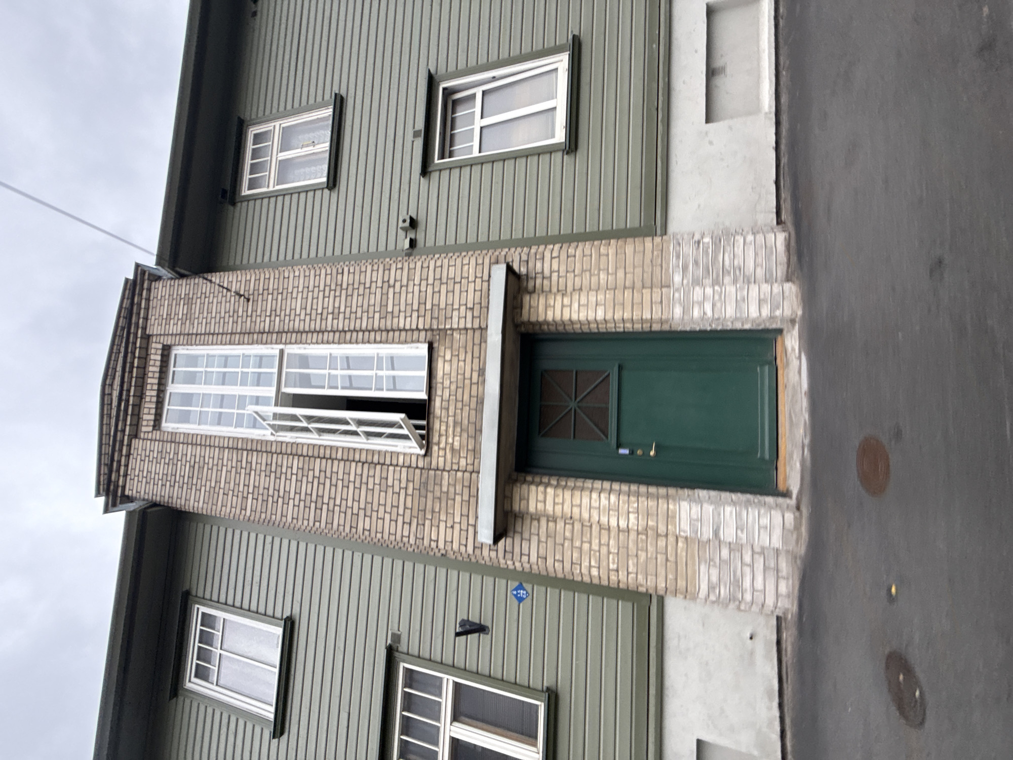 A low-angle, full-shot photograph shows the exterior of a building at Pärnu mnt 41 in Tallinn, Estonia, on a cloudy day. The building features a two-story structure clad in horizontal green siding, with a prominent brick-clad central tower extending upwards. The tower has a tall, narrow window with multiple panes that is partially open. Below this is a dark green door with a geometric pane design and a silver handle. Above the door, a thin metal overhang extends outwards.
To the left of the central tower, a single window with a similar pane design is visible on the upper floor. A small blue diamond-shaped sign with white numbers "3.5 6.8" is affixed to the green siding below this window, next to a black metal fixture. To the right of the central tower, the green siding continues with two more windows visible on the upper floor, also with multi-pane designs. A security camera is mounted on the brickwork to the right of the upper window.
The base of the building, beneath the windows and door, transitions to a lighter gray material. The ground in front of the building is paved with asphalt and shows some circular manhole covers and damp patches. The overall impression is of a weathered, functional urban building.
