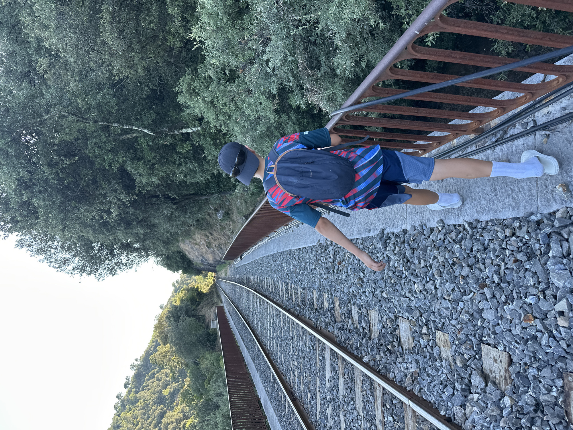 A person wearing a blue, red, and green striped t-shirt, blue shorts, white socks, and white sandals walks along a narrow paved path next to a set of train tracks. They have a dark blue backpack on and wear a dark baseball cap with sunglasses perched on top. The train tracks run parallel to the path and are bordered by a rusty metal railing. The path and tracks are on a bridge or elevated structure. To the left of the tracks, a lush green hillside covered in trees slopes upwards. To the right of the path, dense green foliage and trees are visible. The sky is bright white, suggesting a sunny day. The scene appears to be a scenic natural landscape, possibly in a rural area outside of a city. Specifically, this image was taken near the Cévenol railway viaduct in Anduze, France.