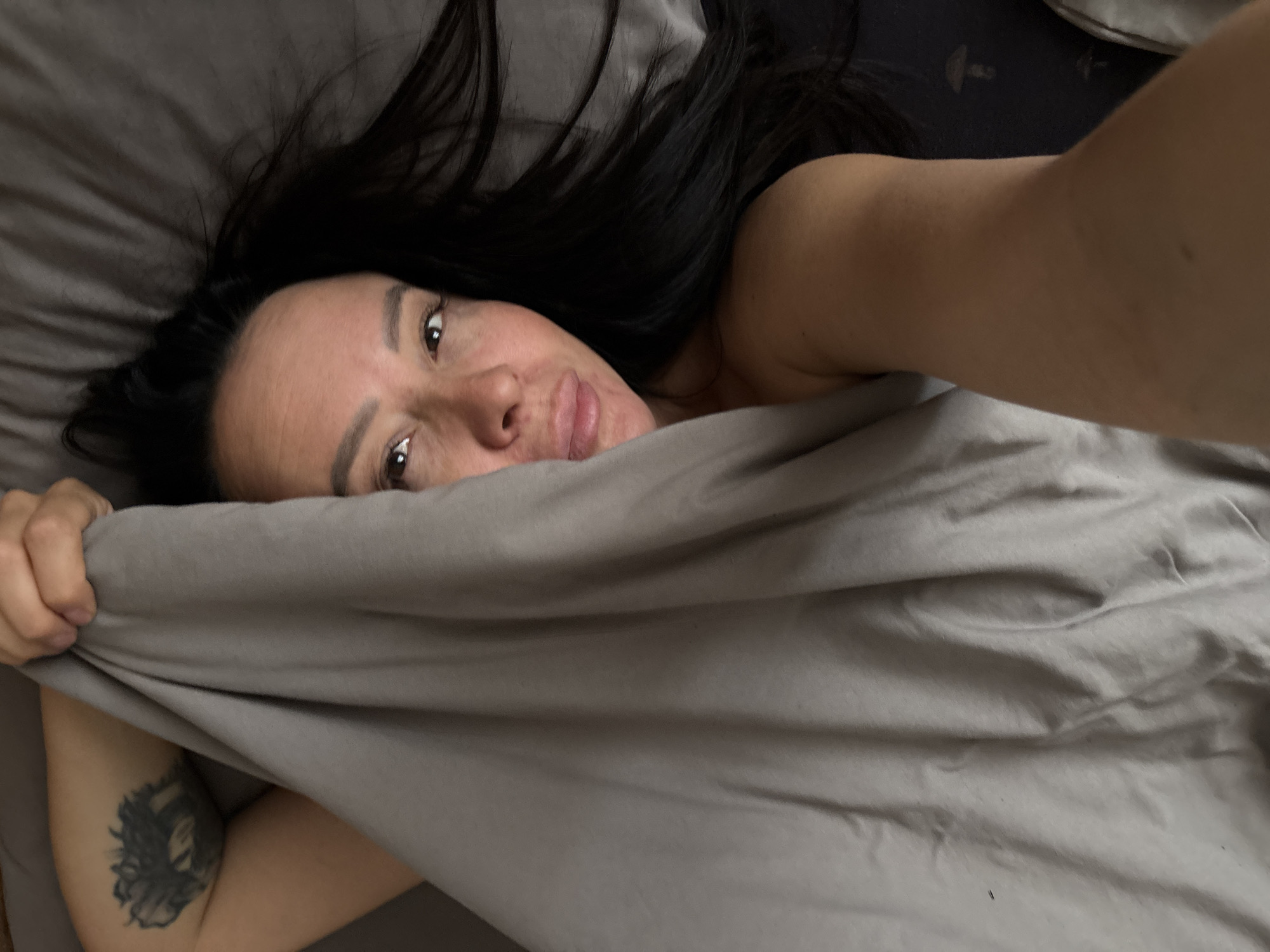 A woman with dark hair lies in a bed in a room with gray bedding. Part of her body is covered by the gray bedsheet, while the upper portion of her face is visible. Her left arm, with a dark tattoo on the upper part, is holding the sheet up. No evidence indicates the location is Chaikovsky, Russia.