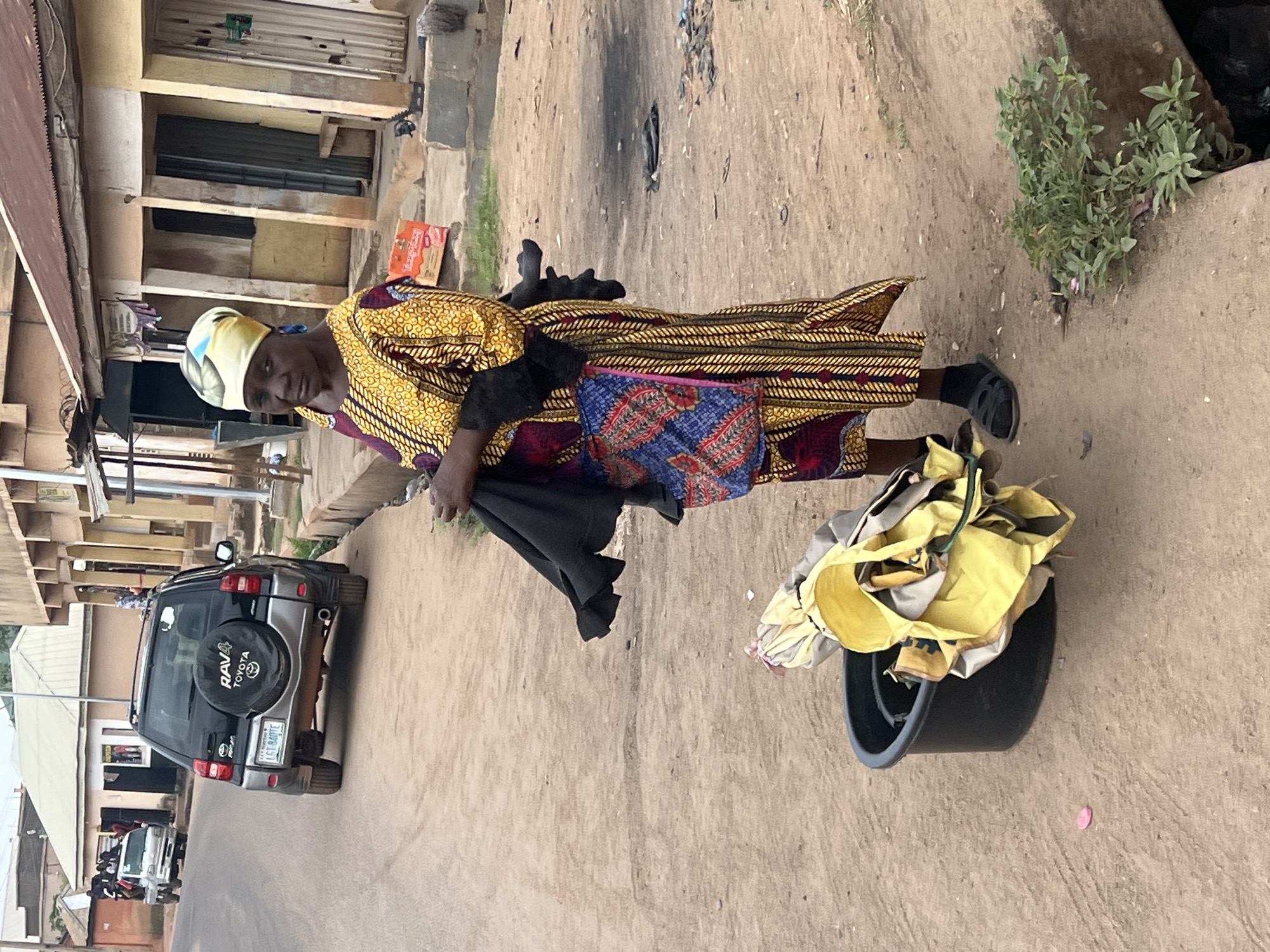 The image, taken in Gura Top, Nigeria, depicts a woman standing on an unpaved road during the daytime. She is dressed in a long, vibrant, patterned gown, primarily in yellow, brown, and red hues, complemented by a light yellow and blue headscarf. She holds a dark piece of cloth in her left hand and looks directly towards the viewer with a neutral expression. Adjacent to her, a black bucket is filled with loosely folded yellow and grey fabrics or clothing items, suggesting she might be a vendor or involved in laundry.
The setting is a local street, characterized by dusty, light-colored ground. To the left of the woman, a grey and black Toyota RAV4 SUV is visible, featuring "RAV4 TOYOTA" prominently on its rear spare tire cover and a Nigerian license plate reading "LGE 840TE". Other vehicles are partially seen further down the road. Buildings line both sides of the street, featuring simple constructions, some with corrugated metal roofs and shuttered windows. Sparse green vegetation grows from the ground near a concrete structure on the right side of the frame. The lighting indicates clear weather conditions, likely morning or afternoon. A small, orange-colored package, with "MAMAS..." partially visible, rests on the ground near the woman.