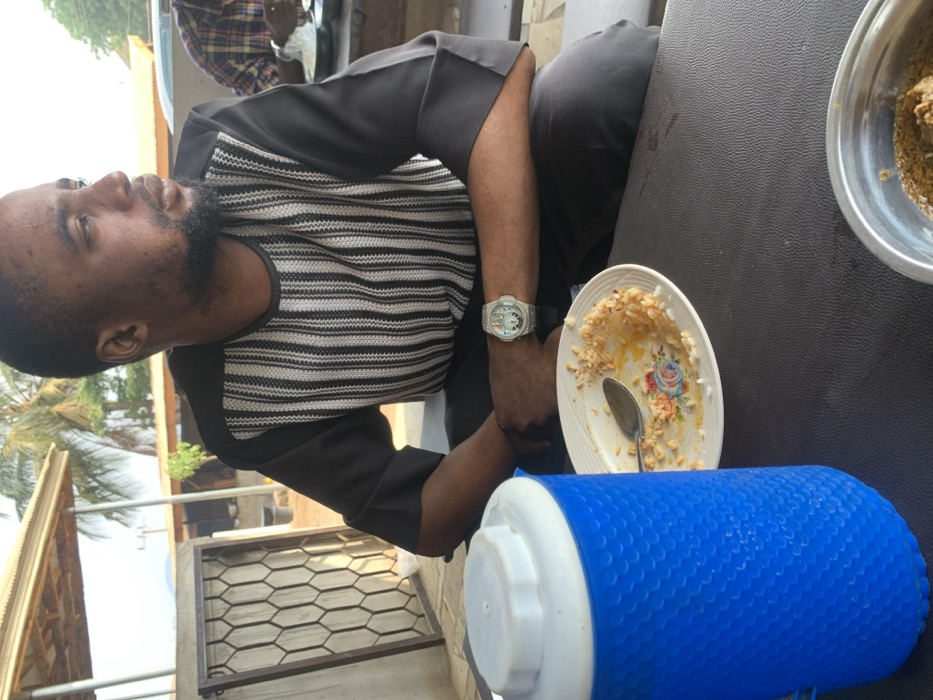A man in a striped shirt sits at a table. He's wearing a watch. On the table are a bowl with food, a plate with remnants of food and a spoon, and a large blue and white water cooler. A building and a palm tree are visible in the background. Another person wearing a plaid shirt is partially visible in the top of the picture.