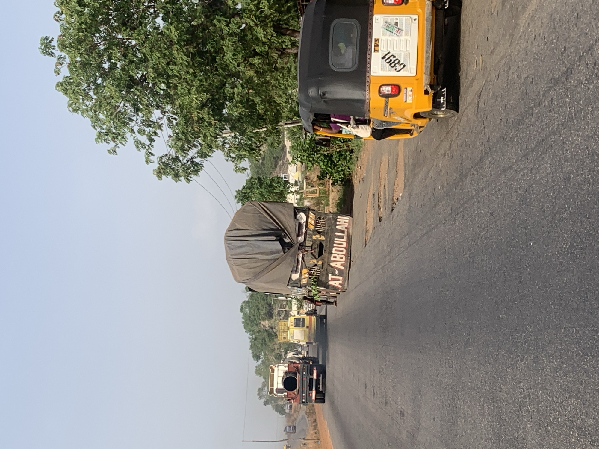The scene is on a road in Lokoja, Nigeria. A yellow, three-wheeled vehicle is parked on the shoulder of the road. A large truck with "AT-ABDULLAHI" written on the back is partially visible behind it. Several other vehicles are farther down the road, including another truck and some smaller vehicles. A tree is on the left side of the frame. The sky is light blue.