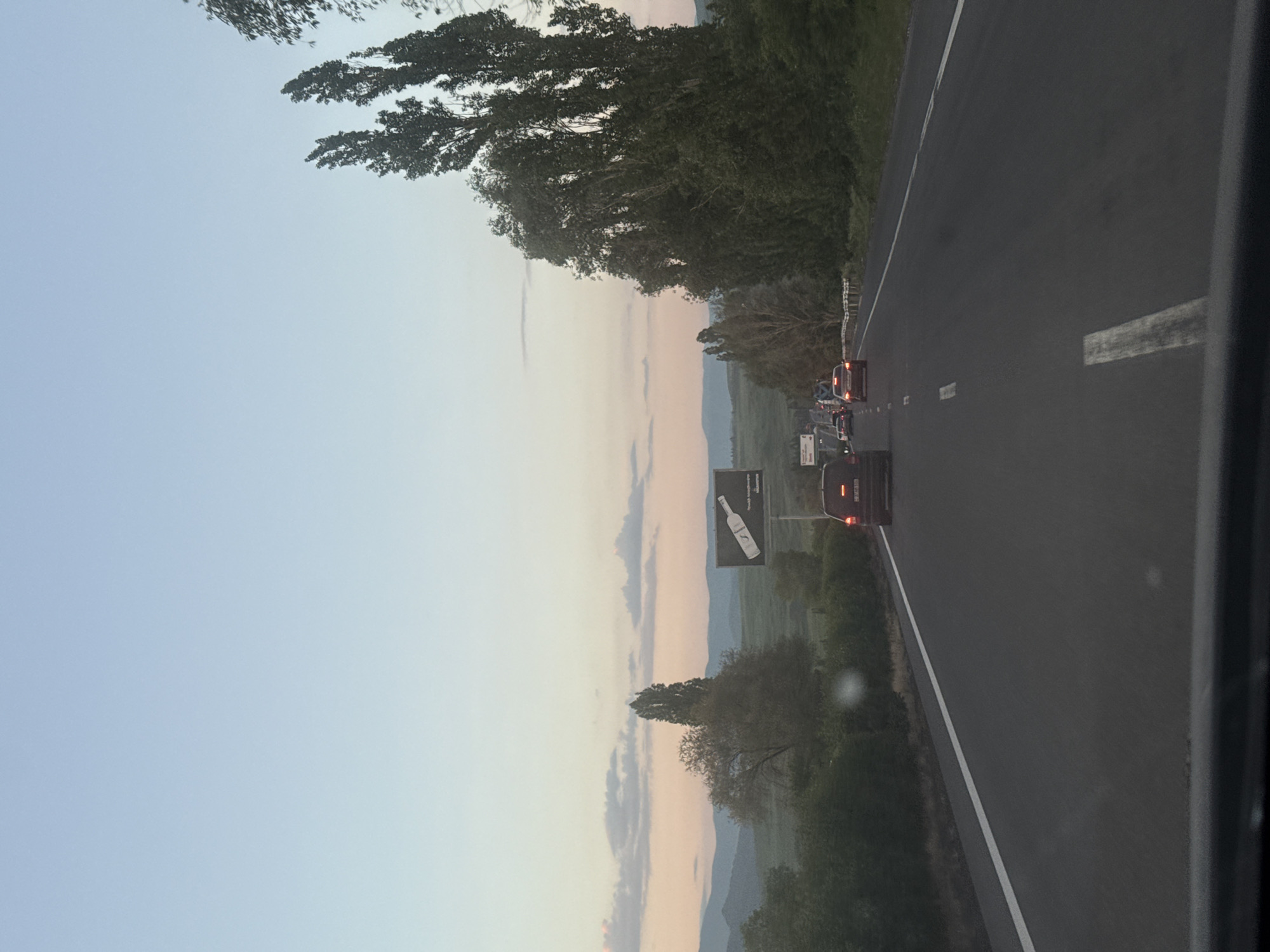 The image shows a road scene at dusk in Armenia. The perspective is from inside a vehicle, looking forward along a multi-lane highway. Traffic is light, with several cars visible ahead. The sky is a soft gradient of pale blue to pink, indicating the sunset. To the right of the road, tall, slender trees line the embankment, their leaves a dark green against the fading light. In the distance, rolling hills are visible, with a billboard featuring an advertisement for a bottled beverage on the left side of the highway. The road surface is dark asphalt with white lane markings. The overall impression is a calm, transitional moment of day.