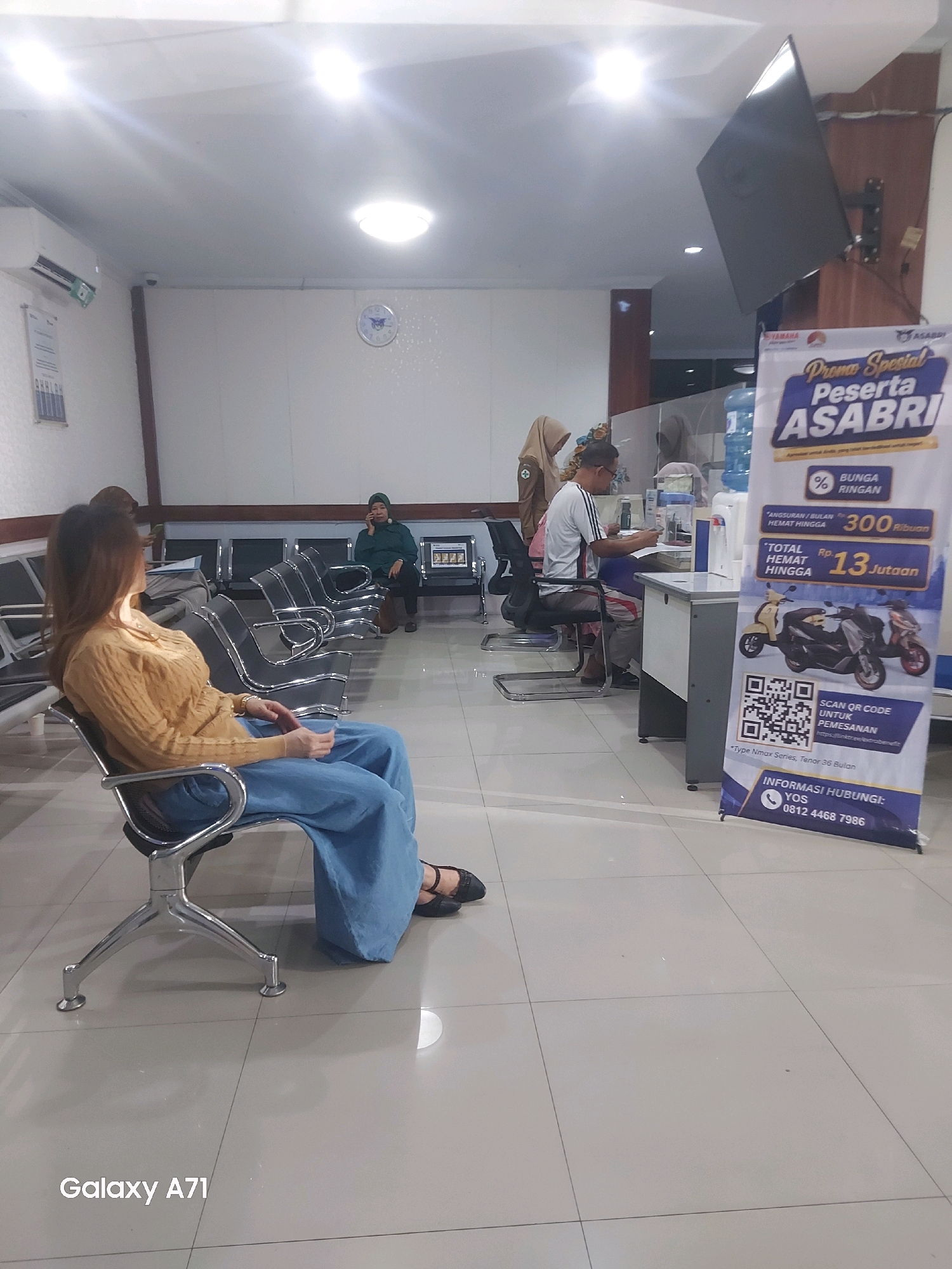 The image depicts the waiting area of what appears to be a government office or service center. In the foreground, a woman with long brown hair, wearing a mustard-yellow knitted sweater and loose blue pants, sits in a metallic chair, facing away from the camera. Her left leg is crossed over her right, and she is wearing dark shoes.
In the mid-ground, several other individuals are present. To the left, a woman wearing a green hijab and a dark green jacket is seated, talking on a cell phone. Behind her, another person wearing a light brown hijab is visible. Further to the right, a man in a white t-shirt with black stripes on the sleeves is seated at a counter, looking down at documents. He is wearing glasses and appears to be interacting with someone off-camera behind the counter.
The environment features a row of empty metallic chairs against a light-colored wall. There is a large promotional banner on the right side of the image, advertising "Promo Spesial Peserta ASABRI" with details about a promotion involving motorcycles. The banner also includes a QR code and a phone number for inquiries. A water cooler is visible behind the counter area. Overhead fluorescent lighting illuminates the scene. On the wall, a clock with a blue face and white hands is visible. An air conditioning unit is mounted on the upper left wall. The floor is tiled in a light, neutral color. The overall impression is that of a public waiting space with a promotional campaign underway. The watermark "Galaxy A71" is present in the lower left corner.