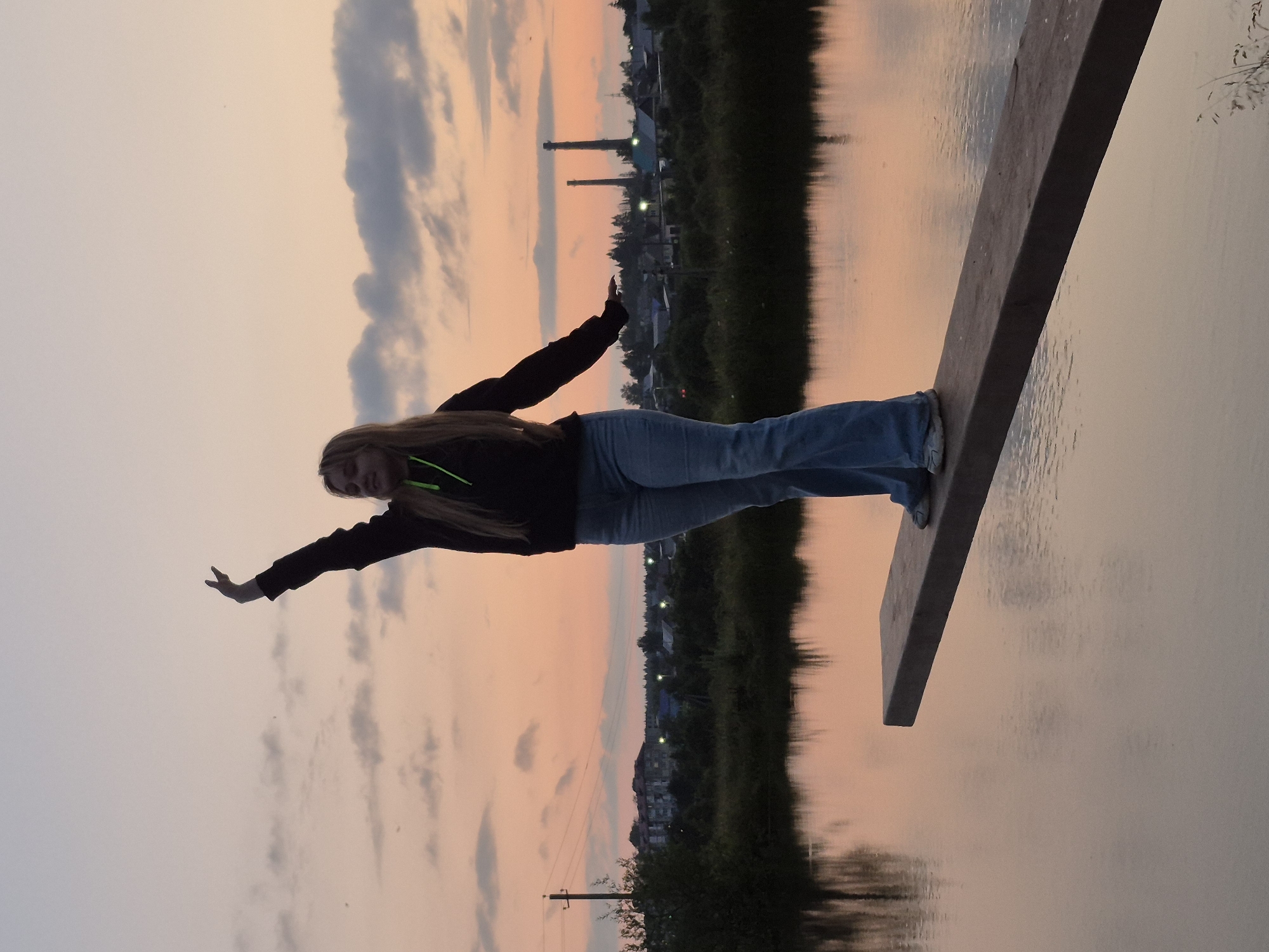 This is a vertical, medium-shot, candid photograph taken at dusk. A young woman with long blonde hair, wearing a dark hoodie and light blue jeans, is captured mid-pose on a wooden plank. Her arms are outstretched, one hand extended forward, the other pointing backward, giving the impression of flight or a superhero pose. She is looking towards the camera with a smile. The plank is positioned over a body of water, which reflects the soft hues of the sunset and the surrounding landscape.
The background depicts a serene scene with a dusky sky transitioning from light grey to soft orange and pink hues. Scattered clouds are visible. Along the far bank of the water, there is a dark line of trees and vegetation, beyond which a village or small town can be seen with houses and lights. Two tall industrial chimneys are prominent in the distance. The water's surface is calm, with gentle ripples.
The time of day is clearly evening, as indicated by the sunset colors in the sky and the reflections on the water. The weather appears calm. The location is identified as Sars, Russia. The overall mood is playful and adventurous, with the young woman expressing joy and freedom in her pose against the backdrop of a peaceful natural setting. There is no visible text in the image.