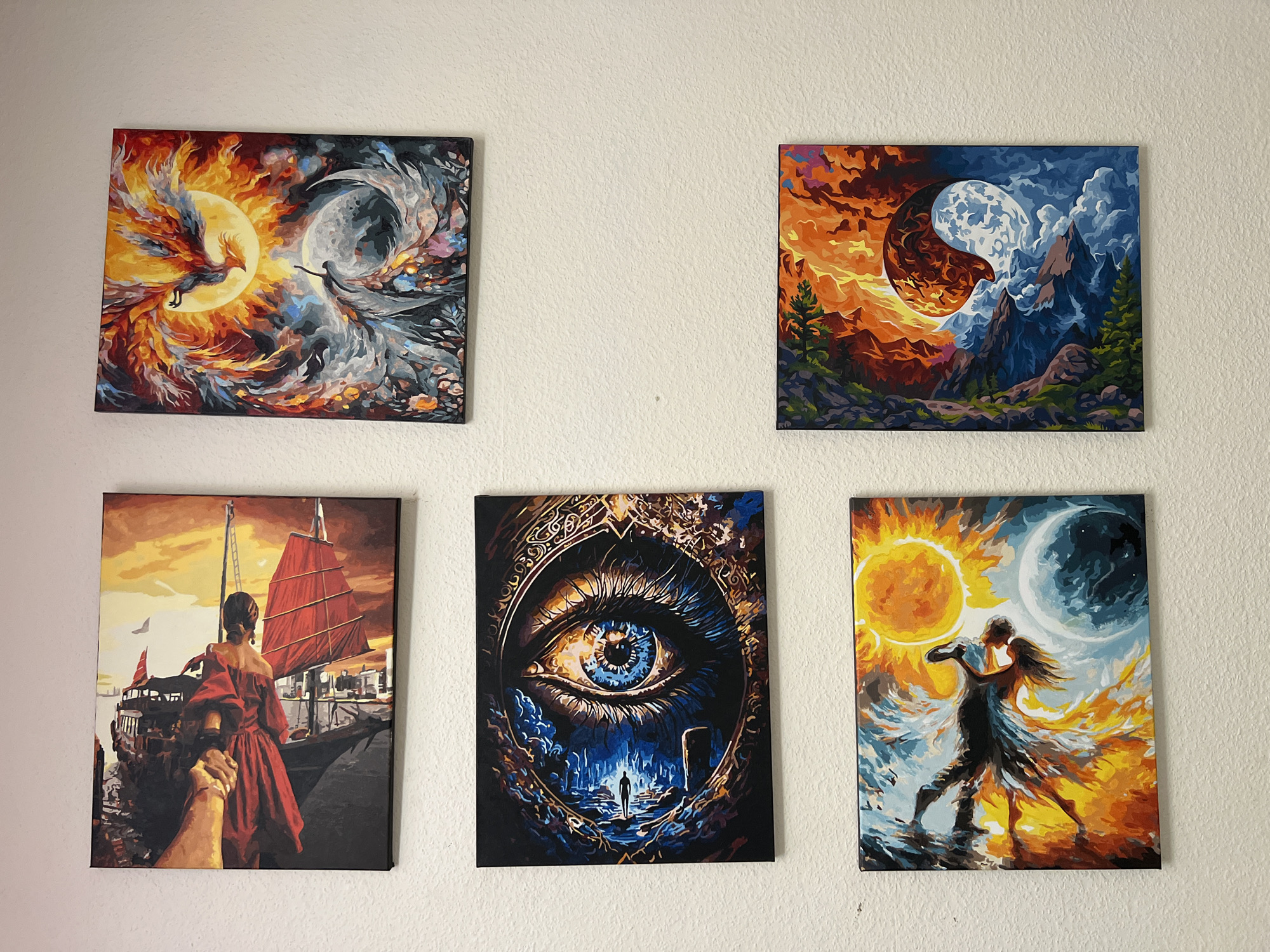 Five framed paintings are displayed on a textured, off-white wall. The paintings are arranged in two rows: the top row has two paintings side-by-side, and the bottom row has three paintings.
The top-left painting depicts a stylized phoenix rising from flames on the left, transitioning into a swirling, moonlit sky with a silhouetted figure on the right. The colors are dominated by fiery oranges, yellows, and cool blues and grays.
The top-right painting shows a landscape divided into two halves by a yin-yang like swirl. The left side features a fiery orange sky with silhouetted trees and mountains, while the right side displays a dark blue sky with a large, detailed moon above rugged mountains.
The bottom-left painting is a scene with a person in a red dress looking away from the viewer, standing next to a large red-sailed ship. The background suggests a harbor with buildings and a sunset sky.
The bottom-middle painting is a close-up of a blue and yellow eye, with a dark, cavernous landscape and a lone silhouetted figure at the bottom. The eye is framed by an ornate, swirling border.
The bottom-right painting shows two silhouetted figures dancing against a backdrop of a large, bright sun on the left and a crescent moon on the right. The background is a vibrant explosion of oranges, yellows, blues, and whites.