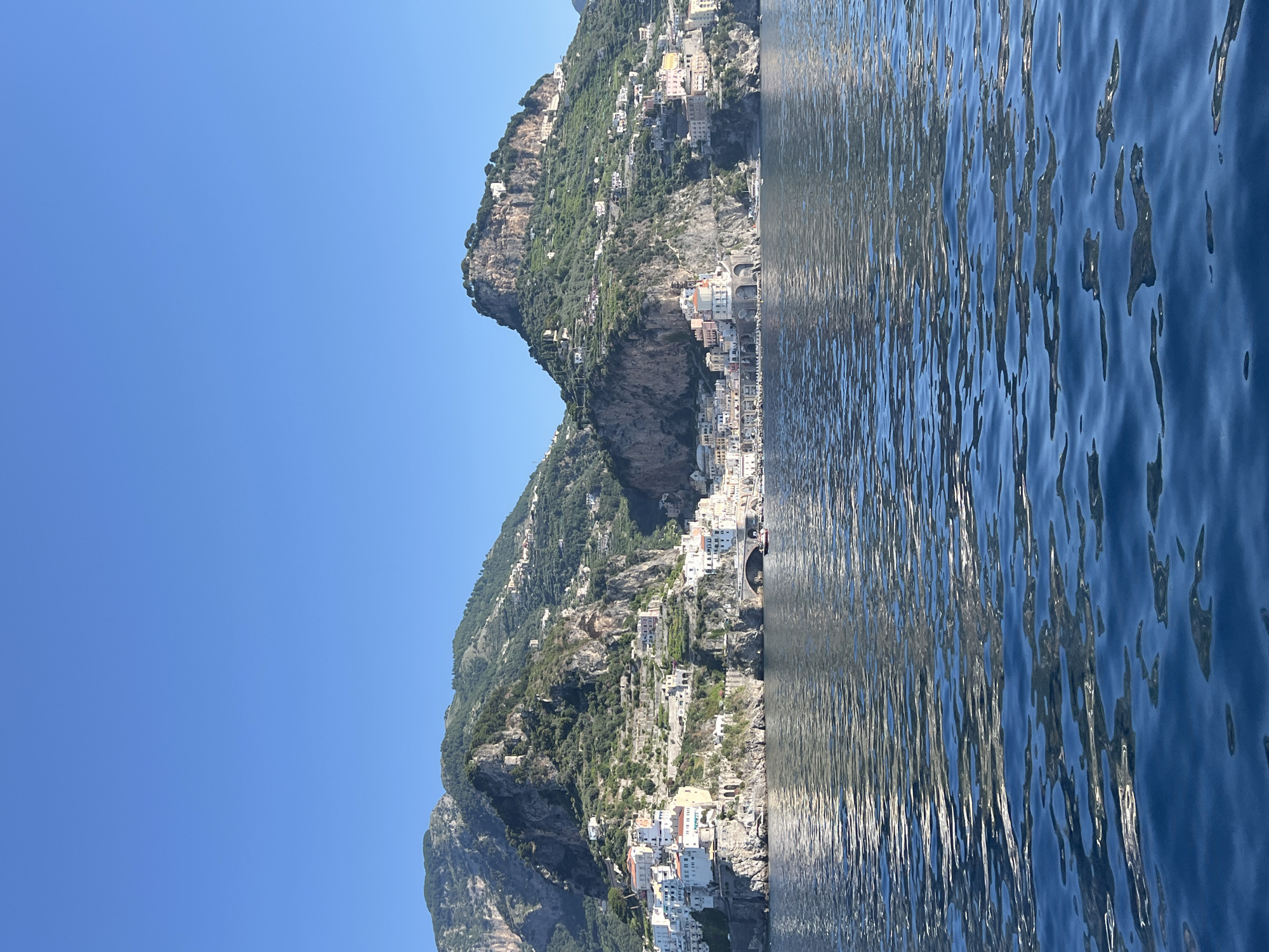 This is a horizontal, eye-level, medium-wide shot of the Amalfi Coast, specifically depicting the village of Atrani nestled at the base of steep, green mountains that rise behind it. The village is characterized by a dense collection of white and pastel-colored buildings with terracotta roofs, clinging to the rocky slopes. A notable feature is a large arched stone bridge spanning a section of the coastline. The water in the foreground is a deep blue with ripples reflecting the sunlight and the colors of the sky and land. Two small boats are visible near the shore, one of which appears to be a red dinghy. The sky is a clear, bright blue with no clouds. The image captures the architectural beauty of the Italian coast and its dramatic natural landscape. The scene is devoid of any identifiable people.