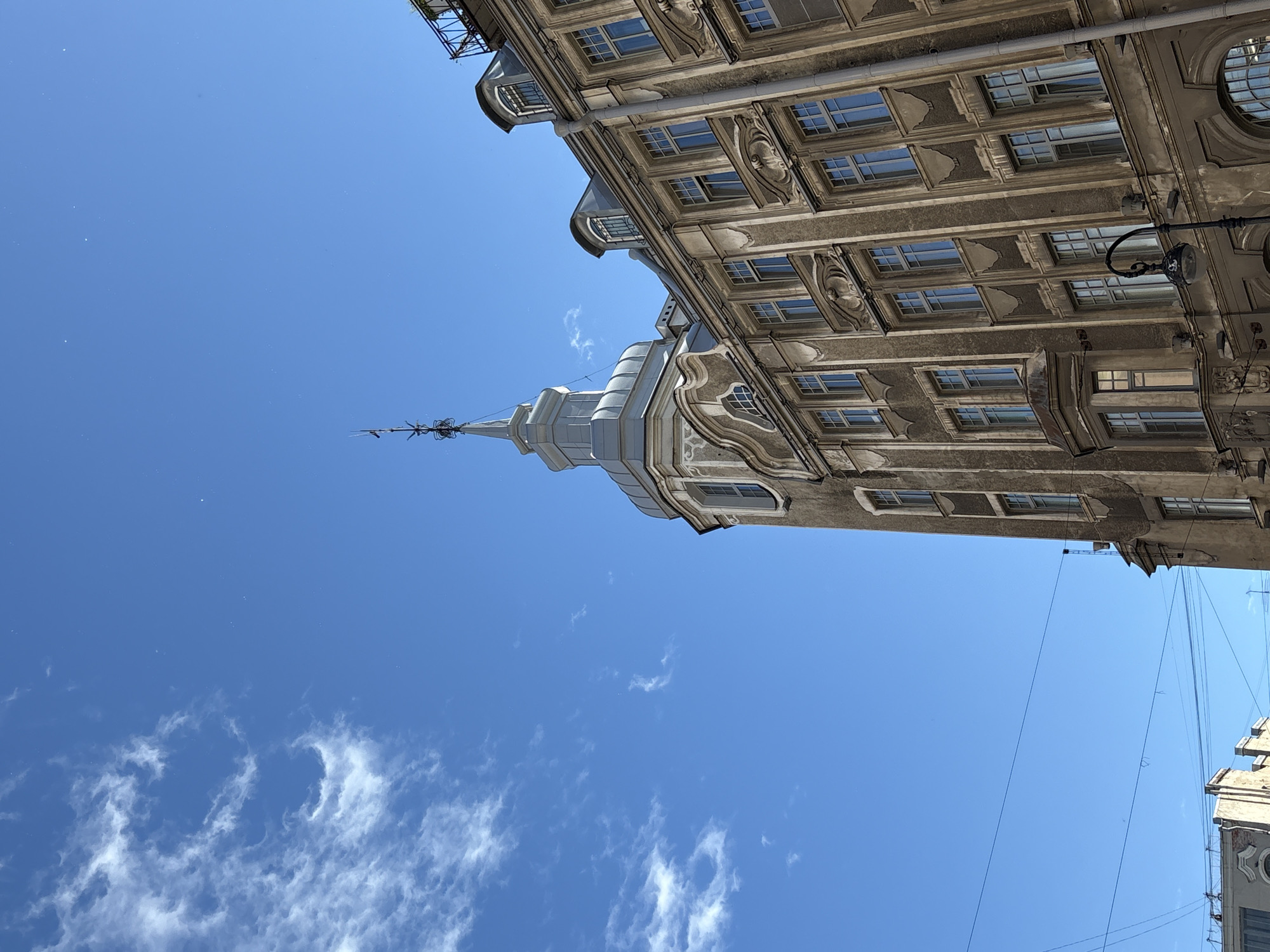 The image depicts an old European building with a prominent turret, set against a bright blue sky with sparse white clouds. The building, located in Saint Petersburg, Russia, features ornate architectural details, including numerous windows with elaborate frames and decorative elements on its facade. A slender spire crowns the turret. The lower portion of the image shows more of the building's exterior and hints at other structures and overhead wires, suggesting an urban environment. The scene is captured from a low angle, emphasizing the scale of the architecture against the sky.