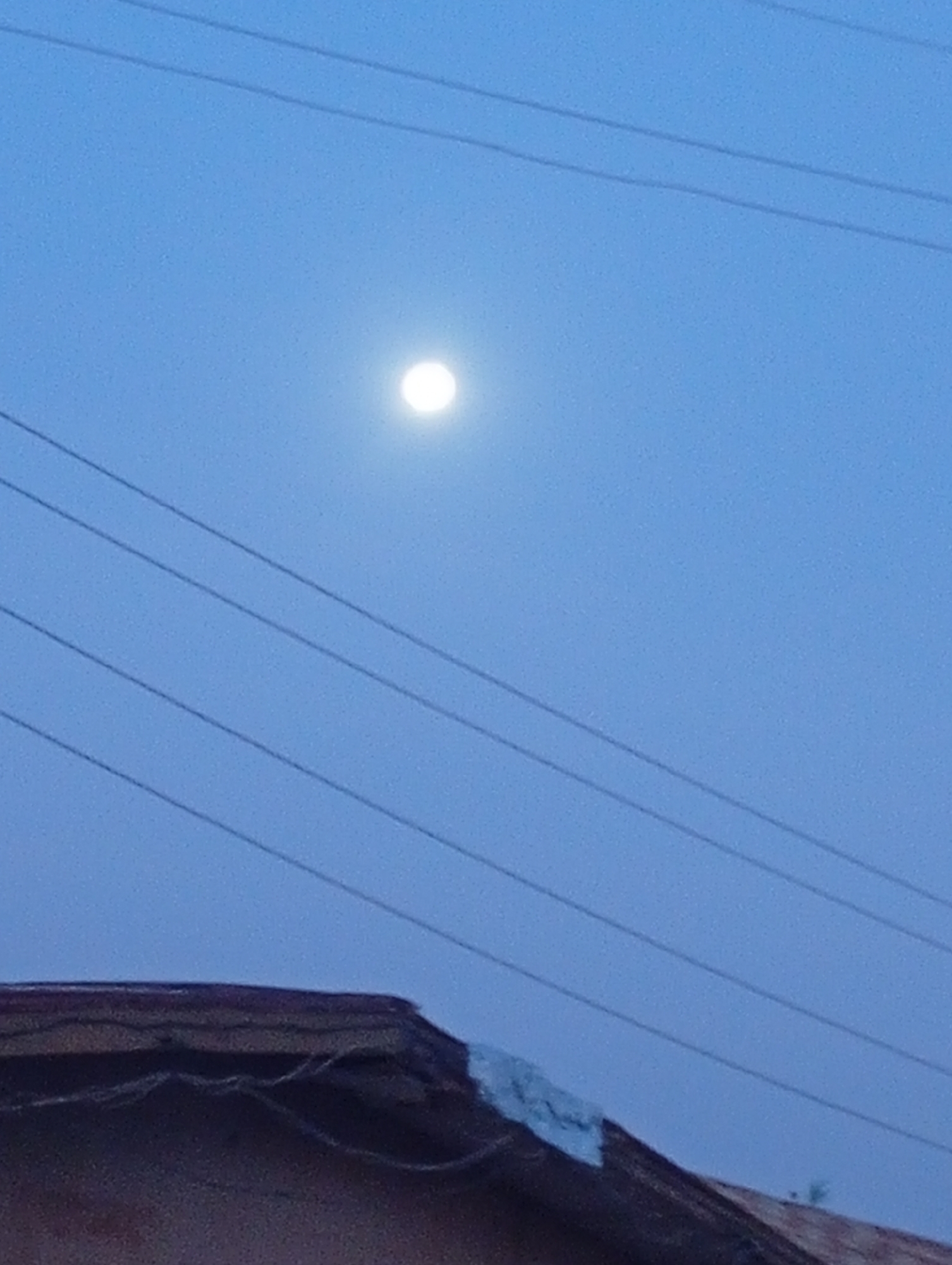 The image shows a night scene in Sabon Tasha, Nigeria. The moon is visible in the upper center of the frame against a dark blue sky. Several power lines stretch horizontally across the sky. In the foreground, the edge of a roof is visible. A patch of silver material appears to be affixed to the roof. The overall lighting suggests twilight or a dimly lit nighttime environment. There are no people visible in the image.