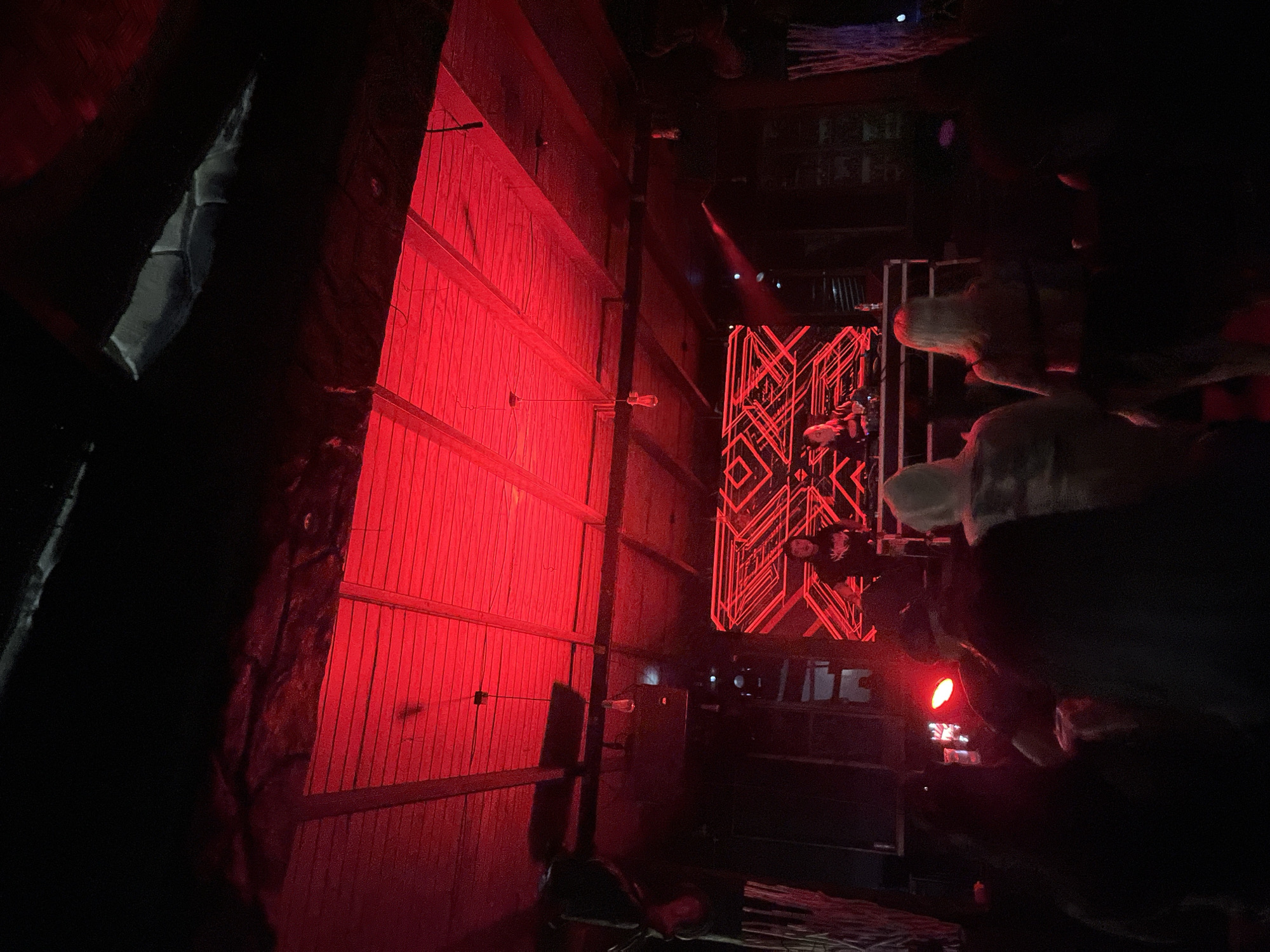 A dimly lit, indoor scene, potentially a performance venue in Arica, Chile, features a vertical wooden panel wall illuminated with red light. A black curtain obscures the left side. A neon sign with a geometric pattern and what appears to be human figures is also lit in red, positioned near a metal cage. A dark, indistinct mass of objects and possible people partially obstructs the right side.