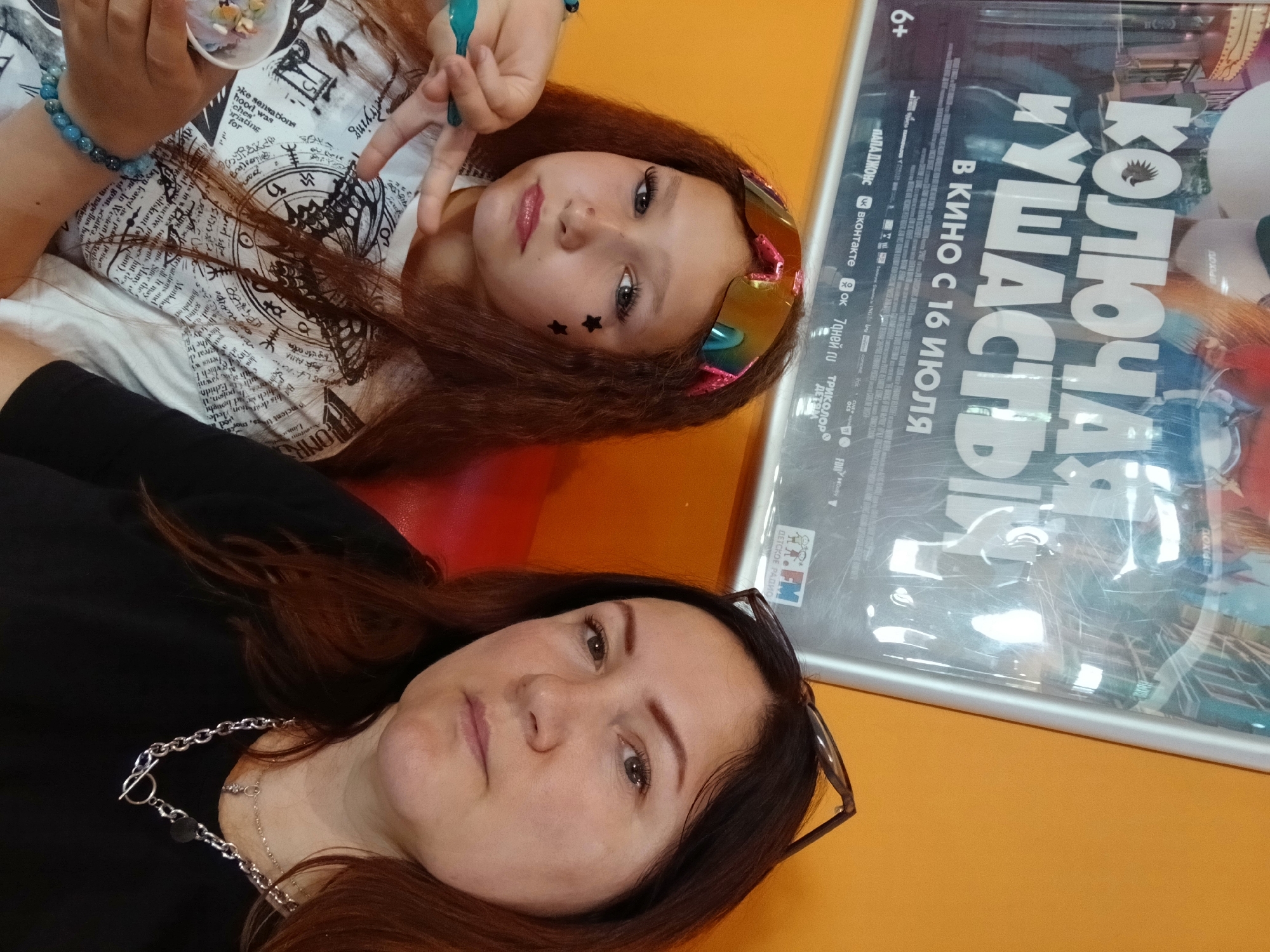 The image is a selfie taken by two females, an adult and a child, likely in a cinema lobby in Chaikovsky, Russia, indicated by the poster in the background advertising a film released on July 16th. The adult, on the right, has dark hair and is wearing a black shirt and a chunky silver necklace. She has glasses perched on her head. The child, on the left, has long, reddish-brown hair and is wearing a white t-shirt with a complex graphic print. She has bright sunglasses with pink frames on her forehead and decorative black stars on her cheek. She is holding a small cup of ice cream or candy and making a peace sign with her fingers. The background shows an orange wall and a large movie poster with Russian text.