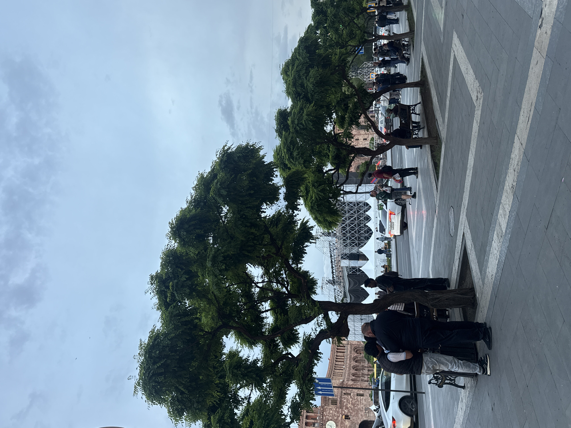 The image captures a scene on a street in Yerevan, Armenia. The foreground features a tree, with people standing nearby. Adjacent is a sidewalk with geometric paving patterns.
Further back, the iconic building of the History Museum of Armenia, with its clock tower, is visible. A stage-like structure is erected behind the museum. The sky is overcast.
People are congregated near the trees and on the sidewalk. Automobiles can be seen parked along the street.