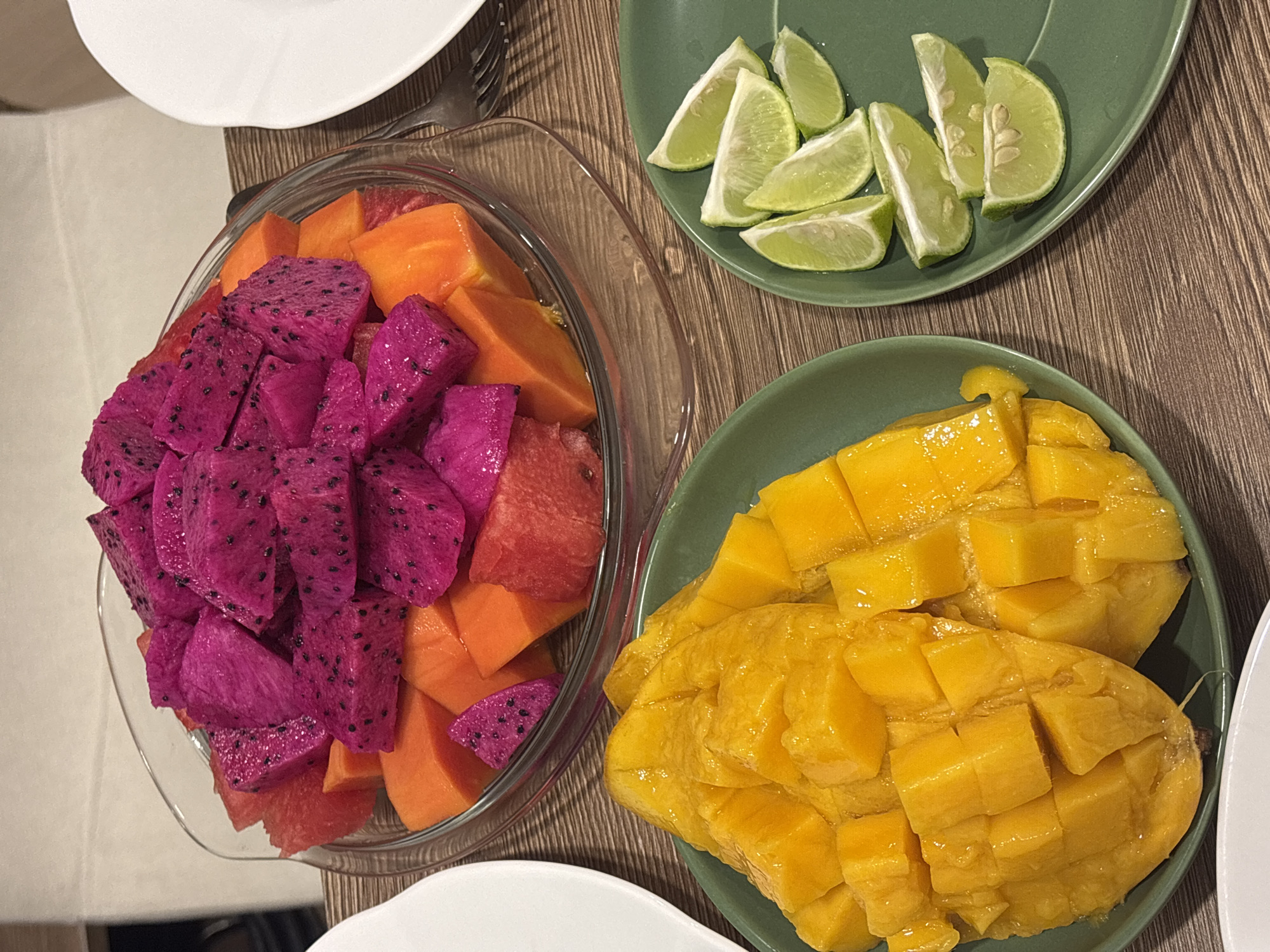 An overhead shot captures a vibrant assortment of fresh, cut tropical fruits laid out on a light brown wooden table. In the center, a large clear glass bowl is filled with a colorful mix of diced fruits, predominantly striking magenta-colored dragon fruit with black seeds, and bright orange papaya cubes. Some red watermelon pieces are also visible within the bowl.
To the right of the fruit bowl, two round green ceramic plates hold additional fruit servings. The upper green plate contains several wedges of fresh lime, some displaying small seeds. Below it, the lower green plate showcases two halves of ripe mango, expertly scored into small, juicy yellow cubes, ready to be eaten. Portions of white ceramic plates or bowls are visible on the top-left and bottom-right edges of the frame, suggesting other place settings. A silver fork is partially seen near the top-left white plate. The wooden table surface has a noticeable grain pattern, contributing to a natural and inviting aesthetic.
The scene depicts a refreshing fruit spread, likely prepared for a meal or snack in a tropical setting, consistent with the location context of Choeng Thale, Thailand. The fruits are presented neatly and invitingly, with vivid colors indicating ripeness and freshness. There is no visible text in the image.