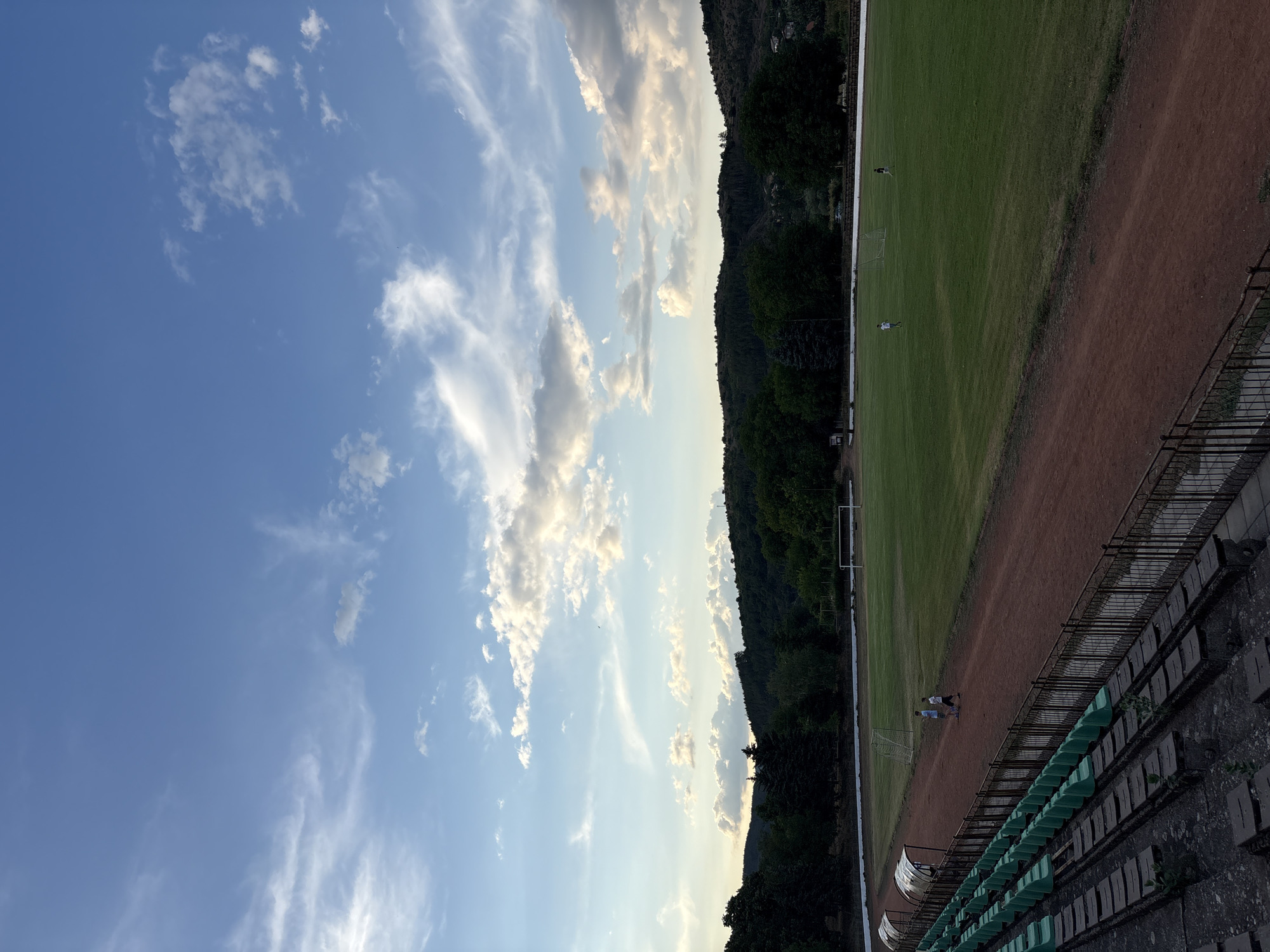 The image shows a view of a sports stadium with a green grass field and a red dirt track. The stadium has rows of empty green seats and some concrete bleachers. Two young boys are walking on the dirt track. In the background, there is a line of trees and hills under a blue sky with scattered white clouds. The location is the stadium in Boboshevo, Bulgaria.