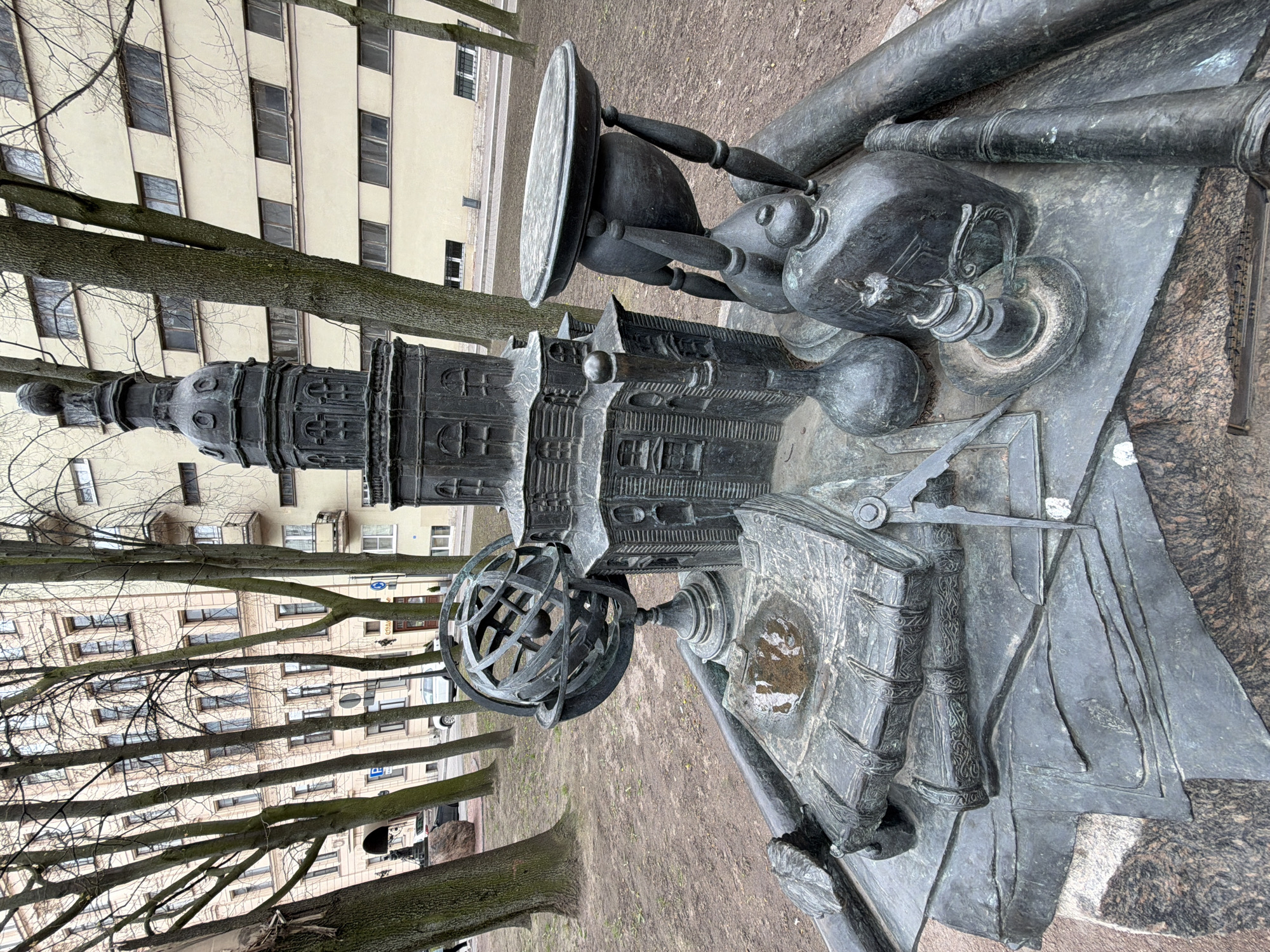 The image captures the "Microbe" monument in Saint Petersburg, Russia. The monument is a bronze sculpture depicting a collection of scientific instruments, including a microscope, books, an hourglass, scrolls, and a globe. These objects are arranged on a stone pedestal. In the background, there is a building and trees. The setting is an urban park or green space, typical of Saint Petersburg. The monument is designed to honor the contributions of scientists and researchers in the field of microbiology.