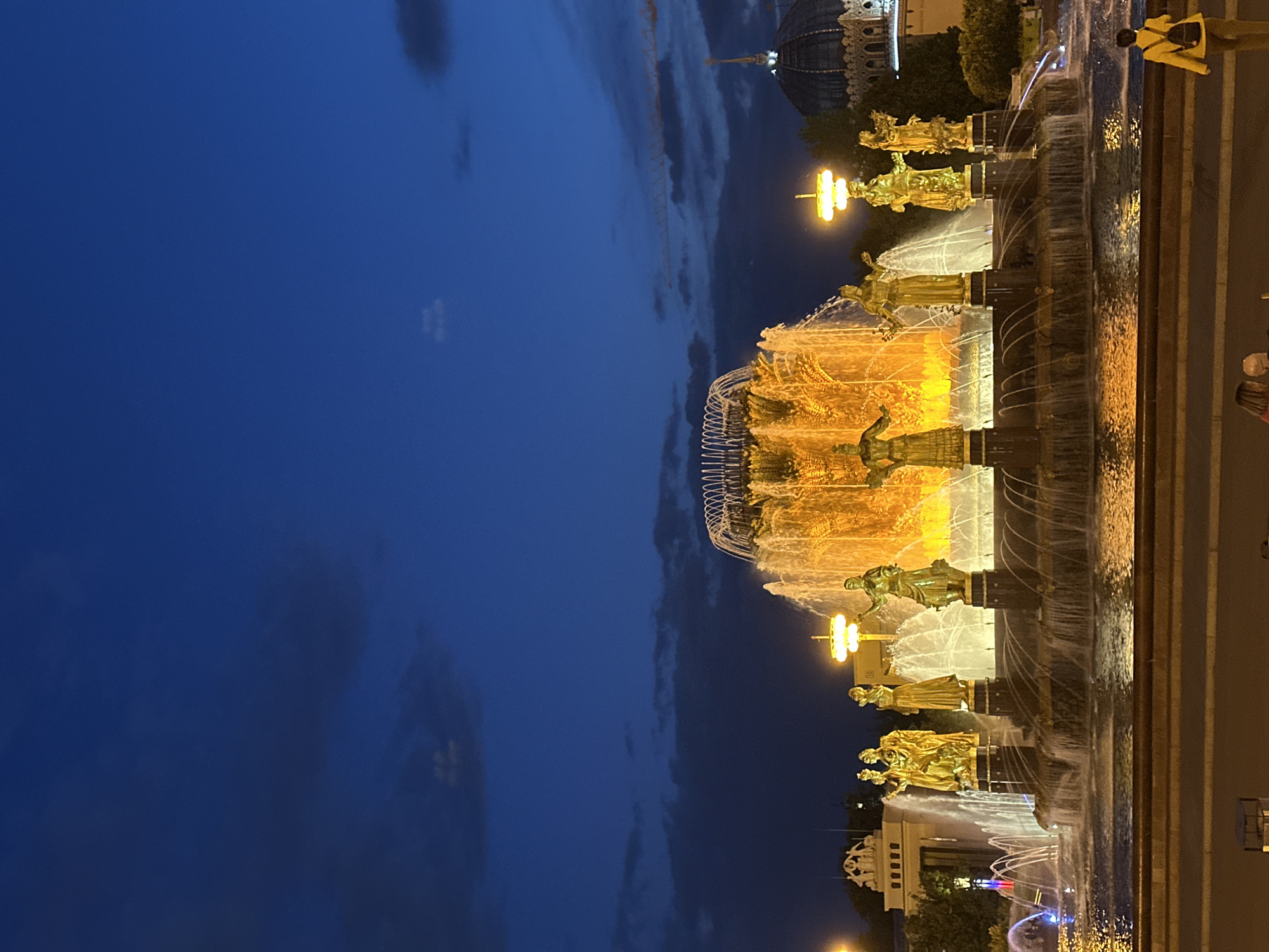 This media file captures the "Friendship of Peoples" Fountain at VDNKh (Exhibition of Achievements of National Economy) in Moscow, Russia, during twilight or night. The prominent subject is the large, multi-tiered fountain, lavishly illuminated with warm, golden lights. This illumination accentuates the numerous golden statues, likely allegorical figures representing the former Soviet republics, positioned around its various levels. Cascading water, also lit by the golden glow, creates a dynamic and radiant display.
In the background, a grand, ornate building with a distinctive dome is visible, showcasing the characteristic Soviet-era architectural style of the VDNKh complex. The sky is a deep, rich blue, indicating the evening hour, with scattered darker clouds adding texture. The foreground features a dark paved area surrounding the fountain, which reflects the bright lights, hinting at either wet surfaces or polished pavement.
Several people are visible in the scene, enjoying the spectacle. One person wearing a yellow jacket is seen from behind, looking towards the illuminated fountain, while another individual in a red top is partially visible. The activity taking place is sightseeing and leisure, with visitors observing the beautifully lit landmark. The overall impression is one of grandeur and serene beauty. No clearly legible text is discernible in the image.
