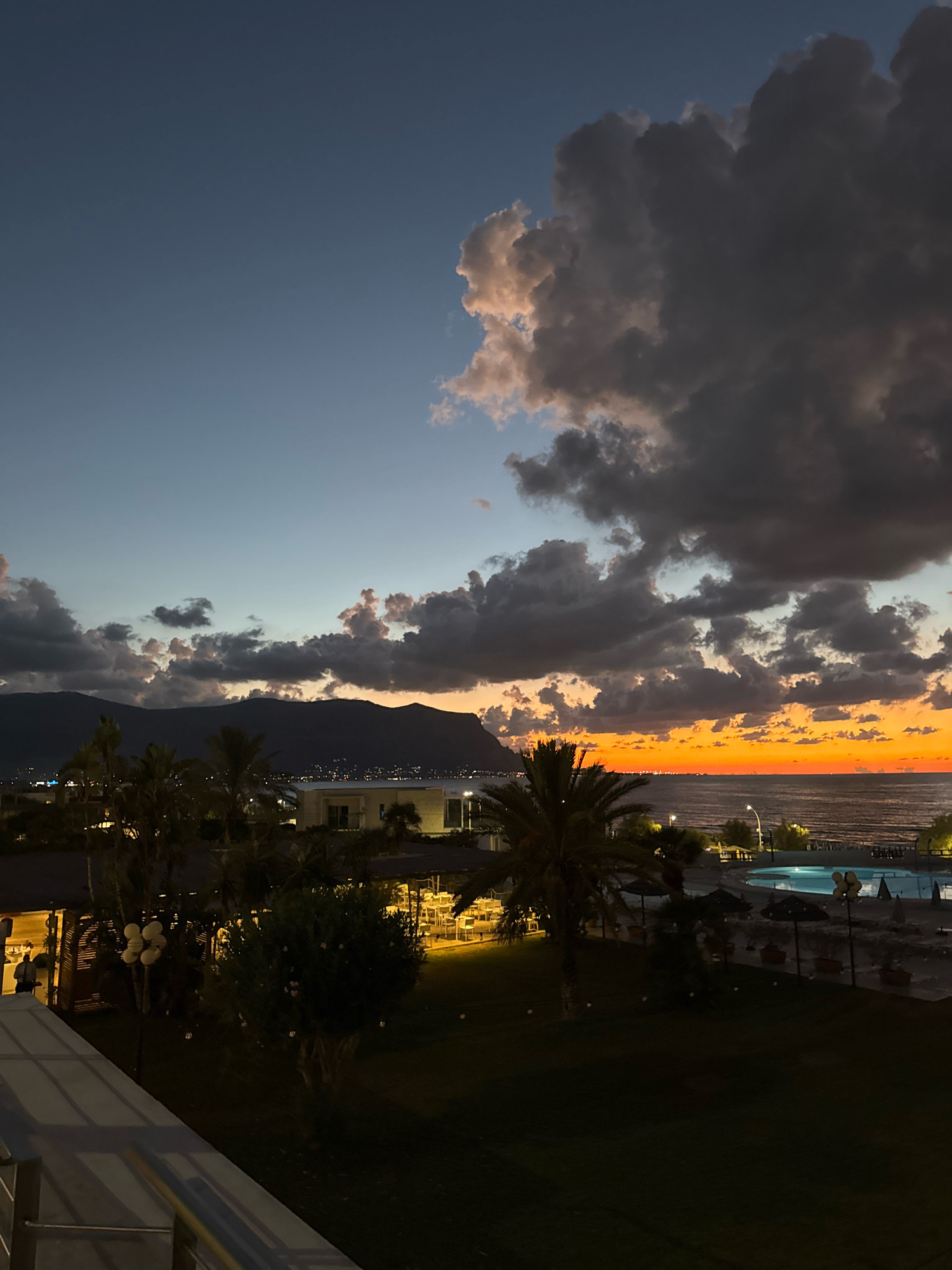 This is a nighttime photograph taken from an elevated vantage point, likely a balcony or terrace, overlooking a coastal area in Isola delle Femmine, Italy. The sky is a gradient of deep blue at the top, transitioning to a soft grey, and then to a vibrant orange band just above the horizon, indicative of sunset or twilight. Dark, dramatic clouds dominate the upper half of the sky, with some illuminated by the receding sunlight.
Below the sky, silhouetted mountains form the backdrop, with faint city lights visible along their base and stretching towards the coast. The sea is visible to the right, appearing dark and calm. In the foreground, the scene is dimly lit, revealing elements of a resort or hotel. There are palm trees, some umbrellas, and outdoor seating areas with tables and chairs, some of which are illuminated by interior lights, casting a warm glow. A swimming pool, faintly lit with blue light, is visible on the right side of the image. A white railing and a portion of a building's exterior are present in the immediate foreground, suggesting the perspective is from a built structure. No people are clearly identifiable, though some figures might be present within the illuminated areas of the buildings. The overall impression is a tranquil, evening scene at a coastal resort.