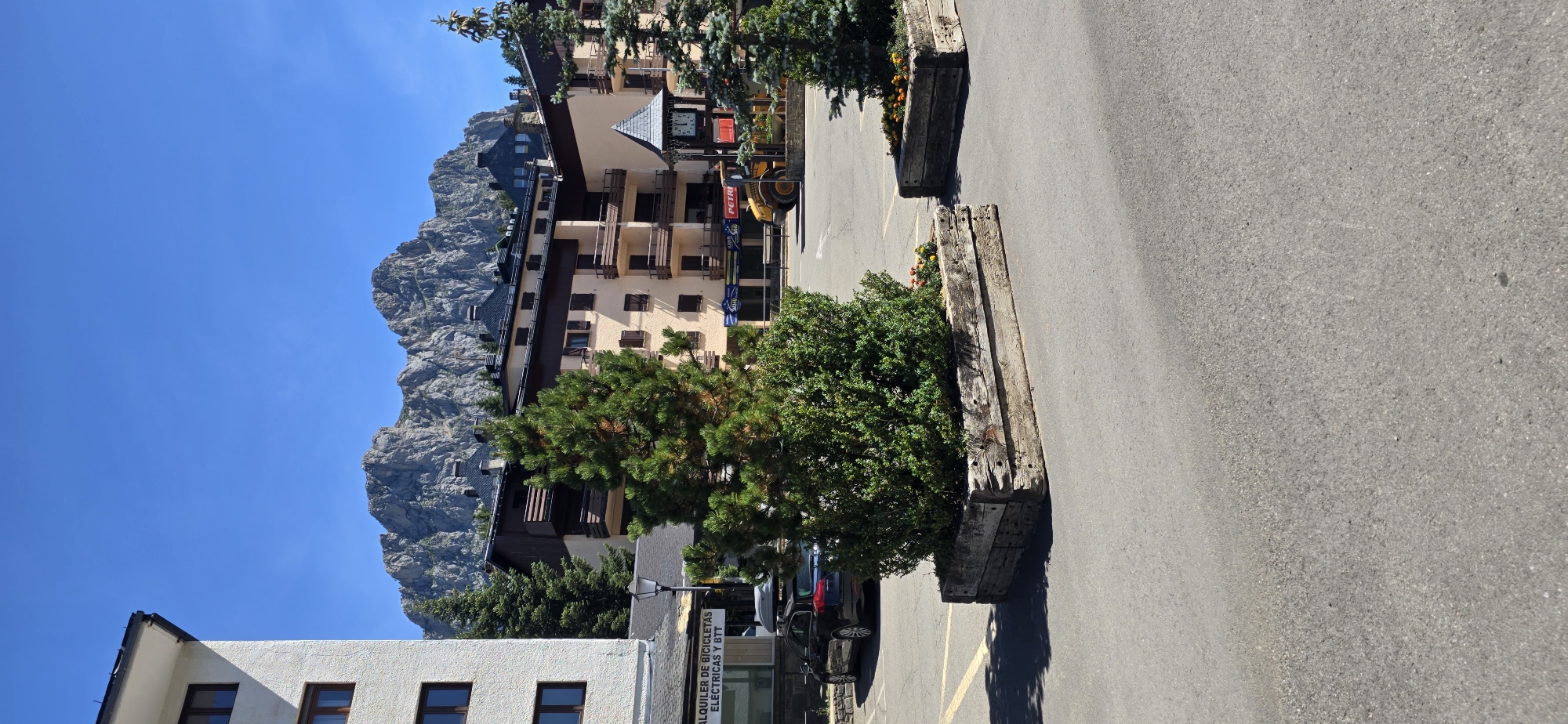 The image shows a view of the town of Sallent de Gállego, Spain, with a focus on the buildings and mountains in the background. In the foreground, there is a paved area with two large wooden planters containing green shrubs and some flowers. A dark colored car with a roof rack is parked on the left side of the frame. Behind the planters, there are several multi-story buildings. The building on the far left is white with dark windows. The central building is light beige with dark brown trim and balconies. To the right of this building is a clock tower. Further in the background, a yellow construction vehicle, likely a loader, is visible. The scene is set against a backdrop of rugged, rocky mountains under a clear blue sky. The overall impression is a sunny day in a mountainous town.