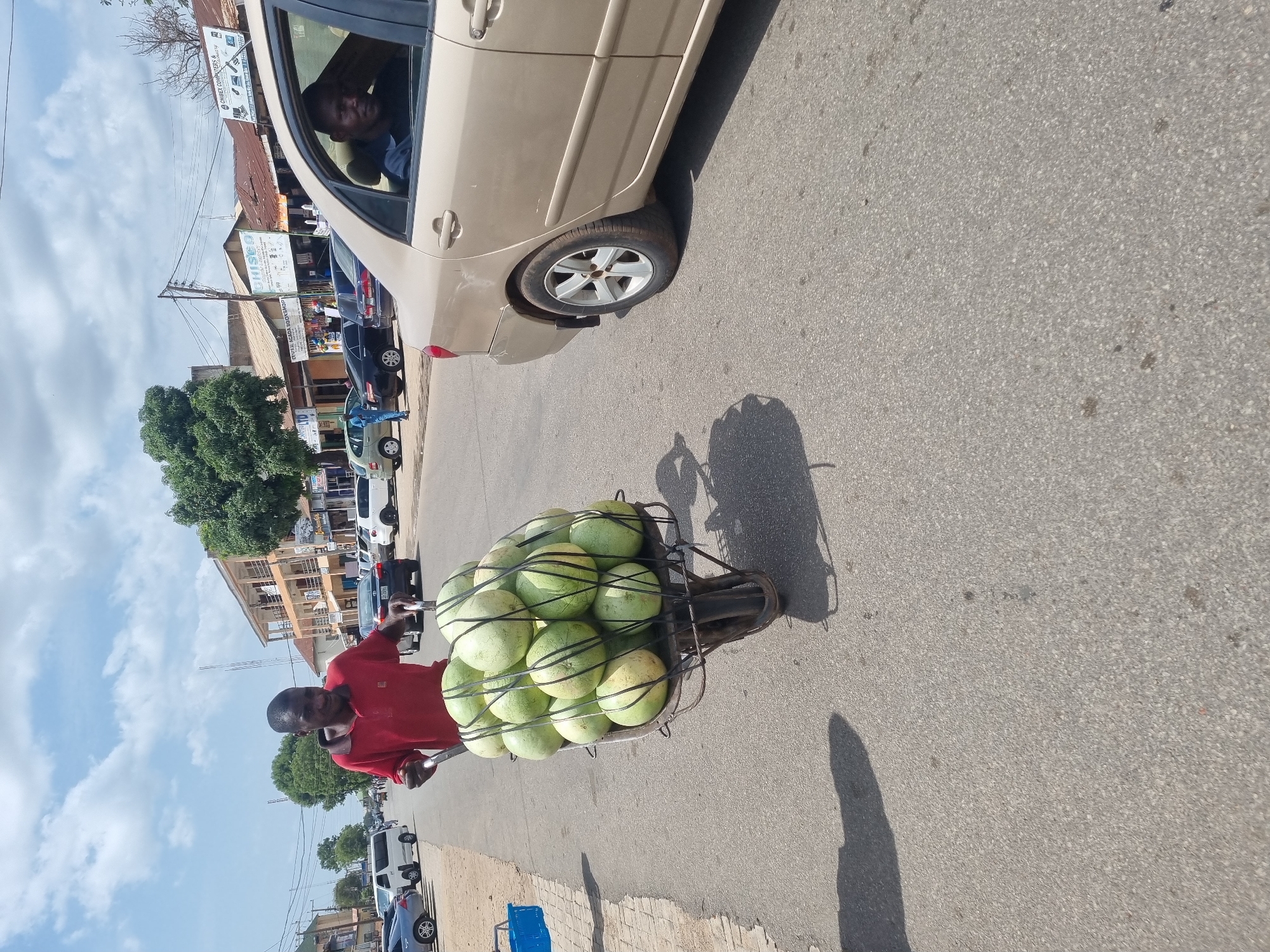 A man wearing a red shirt is on a paved street in Jos, Nigeria, pulling a bicycle cart loaded with watermelons. A tan-colored car is visible on the right edge of the frame, with a man driving the vehicle. Buildings and other parked cars are visible in the background, suggesting an urban setting. The sky is partly cloudy. The street is asphalt.