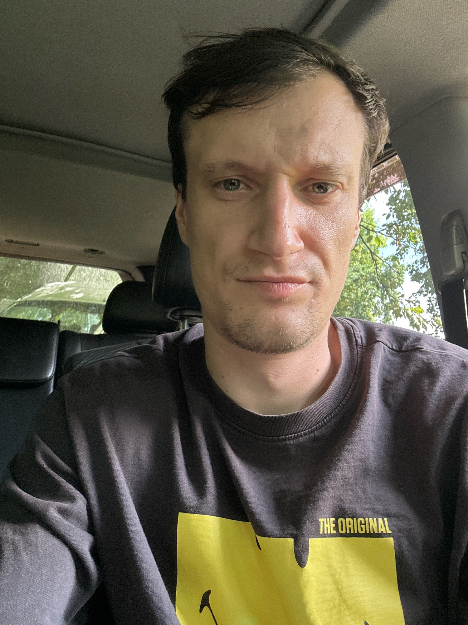A man is taking a selfie inside a vehicle. He is seated in the driver's seat, with the camera pointed towards himself. The man has light skin, short brown hair, and light-colored eyes. He is wearing a dark gray t-shirt with a yellow graphic on the front that reads "THE ORIGINAL". The interior of the vehicle is visible, including the car's roof lining and headrests. Outside the car, trees and foliage can be seen through the windows, suggesting an outdoor setting, possibly a rural or suburban area. The time of day appears to be daytime, with natural light illuminating the scene. The image does not provide specific geographical markers to pinpoint a location within Shatava, Ukraine.