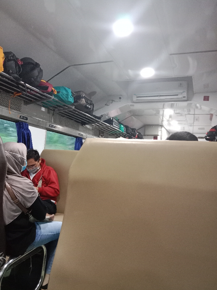 The image shows the interior of a train car in Kedungjati, Indonesia (latitude: -7.178, longitude: 110.664). Two people are seated in the car, a woman wearing a face mask and a gray hijab, and a man wearing a red shirt and a face mask. The woman is sitting on a seat next to the man, and they are both facing forward. The train car is clean and well-lit, with overhead luggage racks filled with bags. The train car has an air conditioner on the ceiling, and there are windows on either side. The car is mostly empty.