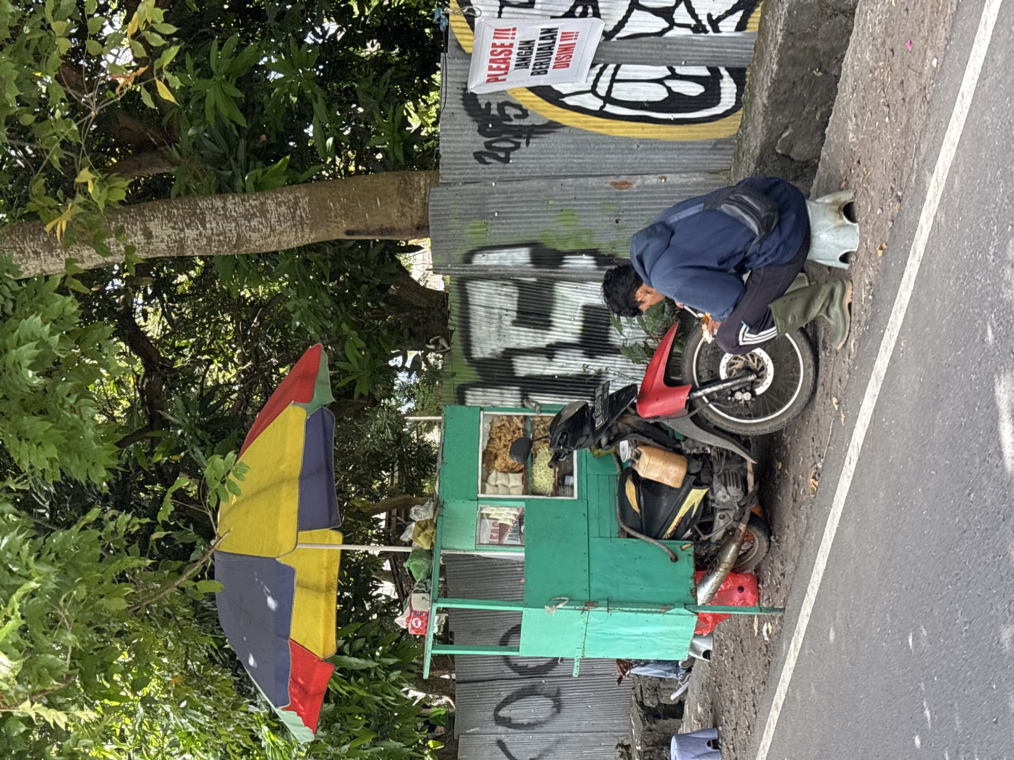 A street scene in North Kuta, Indonesia, features a vendor attending to his mobile food cart. The cart, primarily green with a colorful umbrella attached, is positioned next to a corrugated metal fence adorned with graffiti and a sign reading "PLEASE!!! JANGAN BERJUALAN DISINI!!!". A man wearing a dark blue jacket and dark pants is crouched near the front wheel of a motorcycle connected to the cart. The roadside is adjacent to an asphalt road with a white dividing line. Trees and foliage are visible in the background, suggesting a somewhat vegetated urban setting. The vendor appears to be adjusting or repairing the cart or the motorcycle, indicative of the ongoing maintenance required for a mobile business.