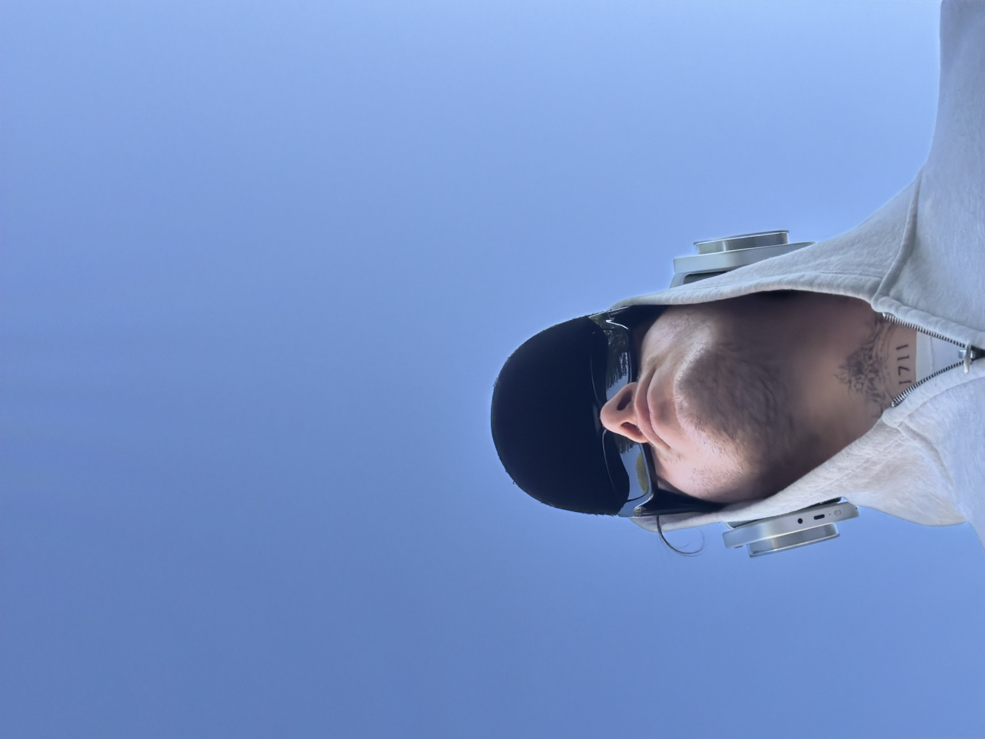 A low-angle, upward-facing shot shows a person with their head tilted slightly to the right. The background is a clear, solid blue sky. The person is wearing a black baseball cap and dark sunglasses. They have light skin and a trimmed beard. They are wearing a light-colored hooded sweatshirt, partially unzipped, revealing a glimpse of a tattoo on their neck with the number "1711". Headphones are visible around their neck. There are no identifiable London landmarks present, and the location is not discernible from the image.
