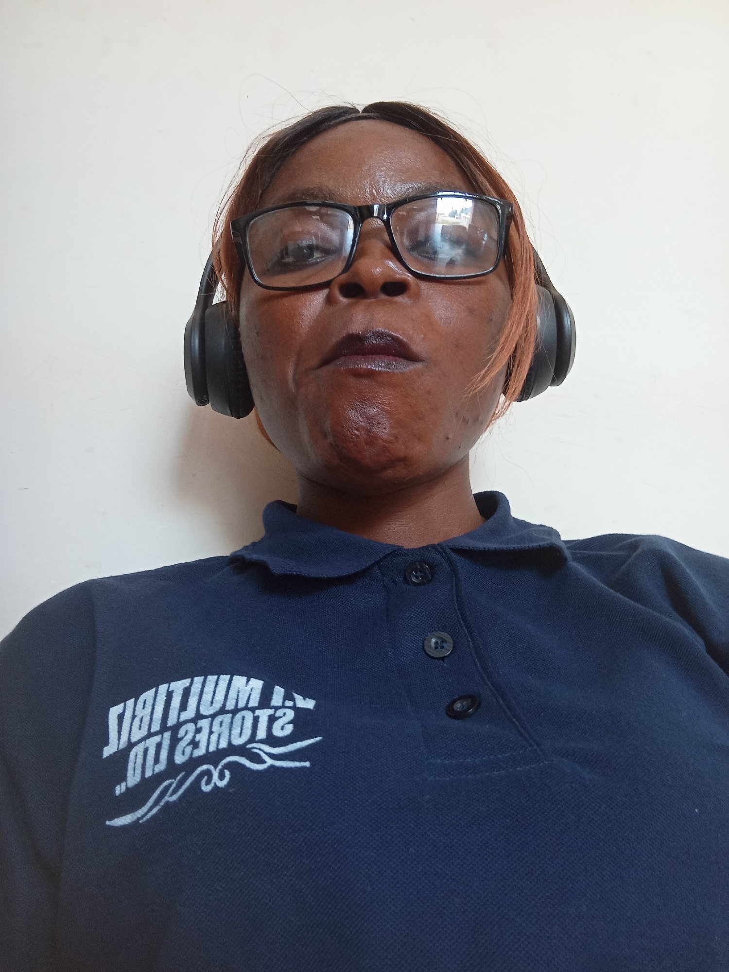 A low-angle, head-on, medium shot captures a Black woman wearing black-rimmed glasses and black headphones. She is looking directly into the camera with her mouth slightly open. Her dark hair has reddish-brown highlights. She is wearing a dark blue polo shirt with two dark buttons on the placket. A white, mirrored logo is visible on the left side of her chest. The background is a plain, light-colored wall. The image appears to have been taken indoors.