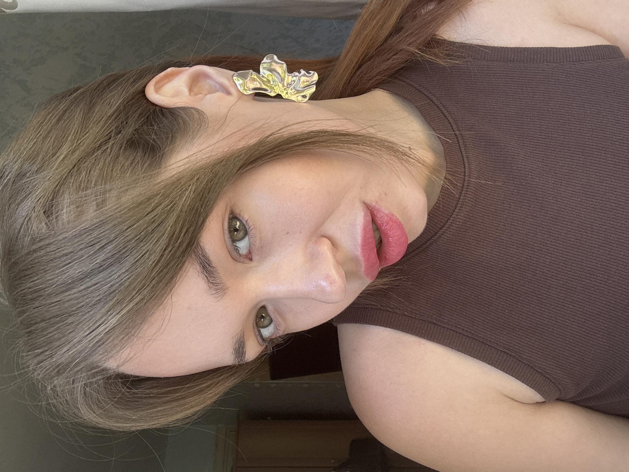 A close-up, outdoor, natural light portrait of a young woman with light brown hair, green eyes, and pink lipstick. She is wearing a brown ribbed tank top and a large, gold, abstract butterfly-shaped earring on her right ear. The background shows a slightly out-of-focus interior space with a dark wooden cabinet and a window with light filtering through, suggesting a residential setting. The image does not provide specific visual cues to pinpoint its location within the city of Chaikovsky, Russia.
