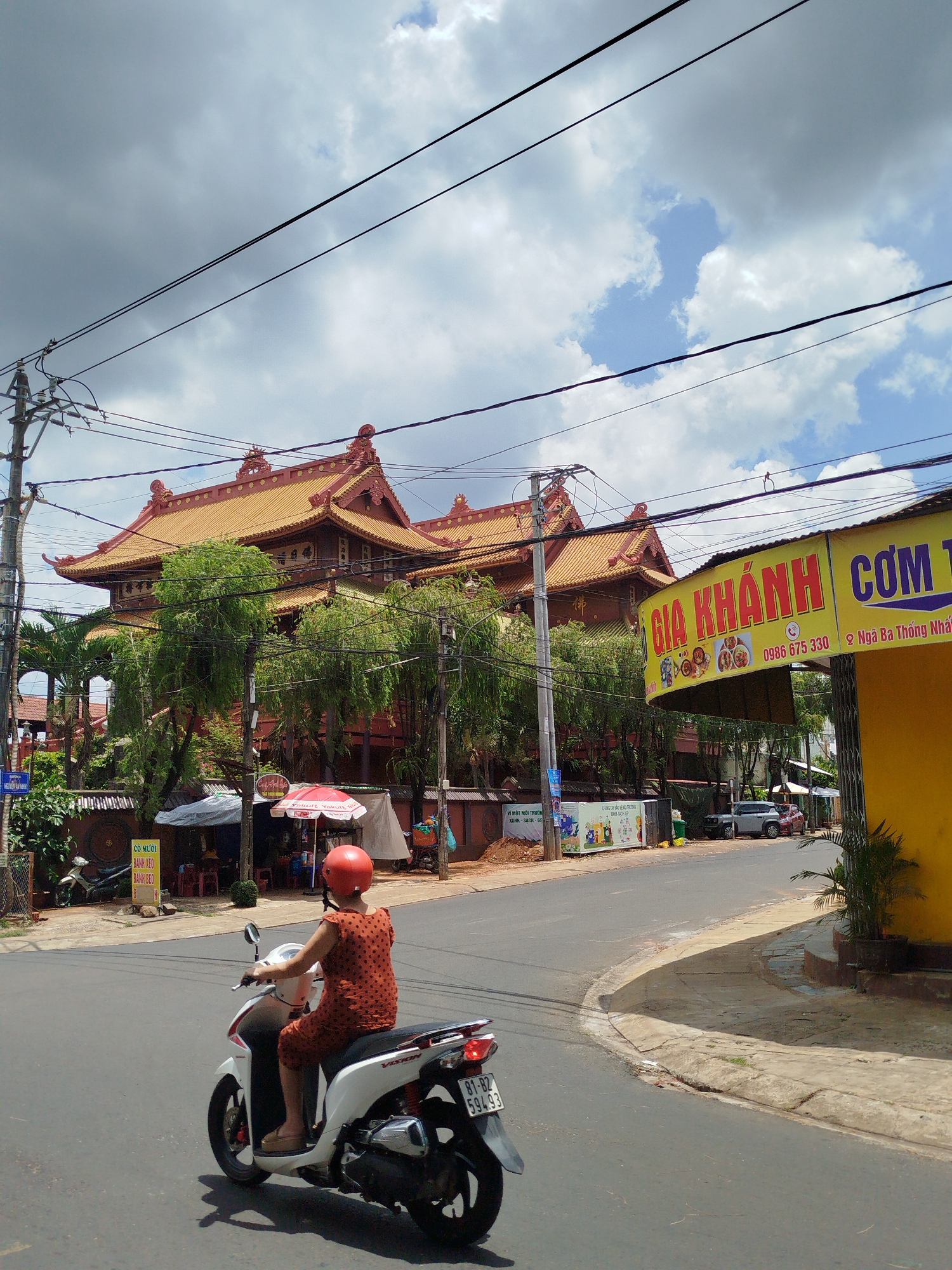 A vibrant, medium-shot captures a street scene in Dien Hong Ward, Vietnam. A woman wearing a red dress and helmet rides a white scooter down a grey asphalt road. In the background, a traditional red and yellow tiered building stands behind a canopy of green trees. A bright yellow commercial sign reading "GIA KHÁNH CƠM T" in red letters hangs on the right side of the frame. Utility poles and wires crisscross the sky above. The sky itself is a mix of blue and grey clouds. Smaller vendor stalls are visible along the roadside near the building. The scooter's license plate is visible: "81-B2 594.93". Other signage is evident, including a sign for "CÔ MƯỜI" and "Yakult Yakult." A light-colored car is parked on the opposite side of the street. Construction or renovation appears to be taking place near the tiered building.