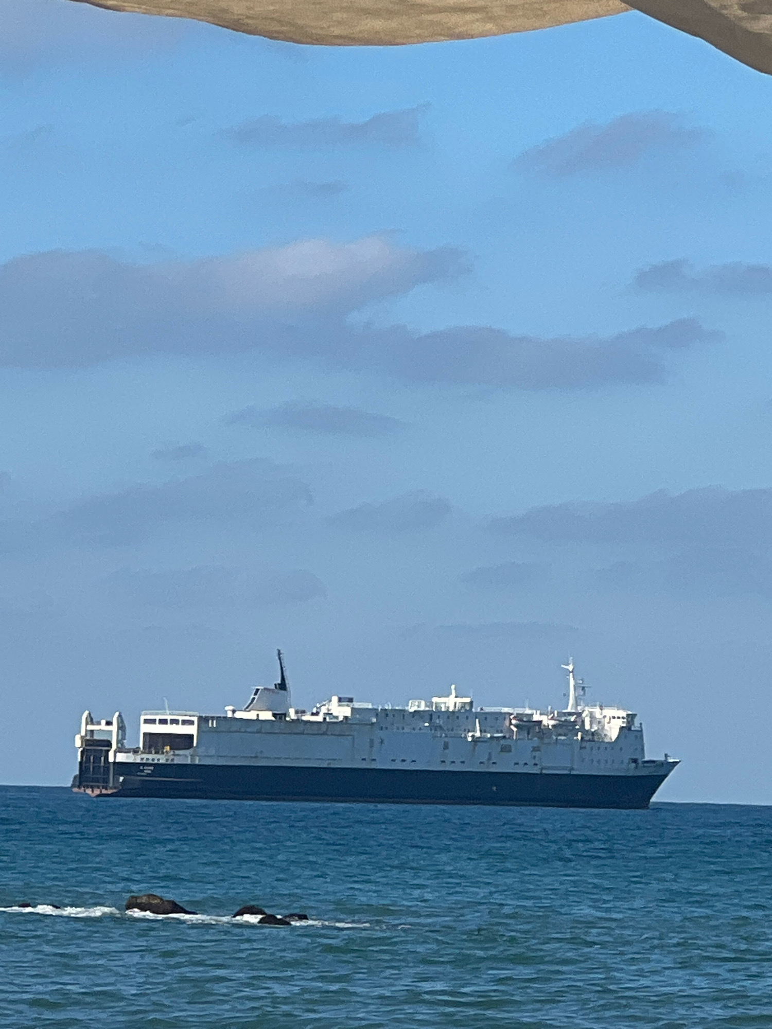 A large cargo ship is sailing on the blue sea under a sky with scattered clouds. The ship is white with a dark blue hull and has multiple decks and structures, including a prominent smokestack. In the foreground, several dark rocks are visible partially submerged in the water, with small waves breaking around them. The scene is likely the coast of Karasu, Turkey, given the context.