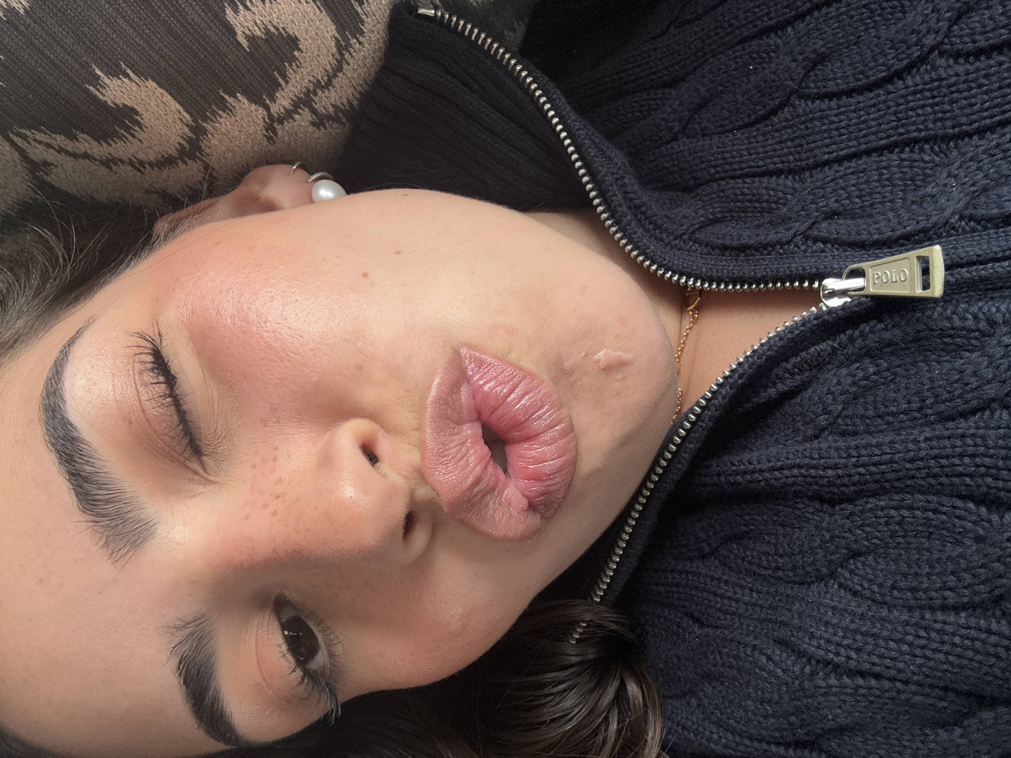 A person is depicted in a close-up photograph. They are lying down, facing upwards at an angle, with one eye closed. The person's lips are pursed as if blowing a kiss. They have dark eyebrows, eyelashes, and hair. Freckles are visible on their face. They are wearing a dark blue, cable-knit, zippered sweater with the brand "POLO" visible on the zipper pull. A gold chain necklace is partially visible at their neck. A ring with a pearl is visible on their hand near the upper left corner of the image. A patterned cushion is behind their head. The location is not possible to determine based on the image provided.