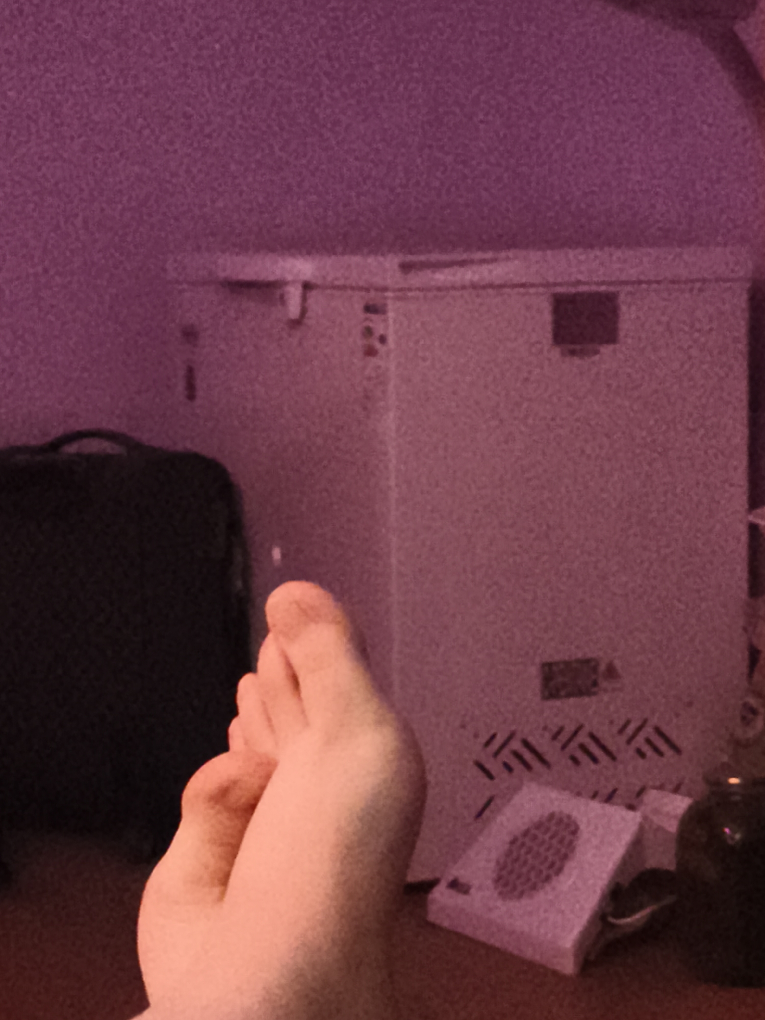 The image depicts an indoor scene, possibly a residential room in Ekibastuz, Kazakhstan. In the foreground, a person's bare foot is visible, resting on a reddish-brown surface. Behind the foot, a dark-colored suitcase or bag is positioned to the left. To the right of the suitcase, a cream-colored appliance with vents is visible. The background is dominated by a wall with a textured, purplish hue. The lighting appears to be dim, with a pinkish cast over the scene.