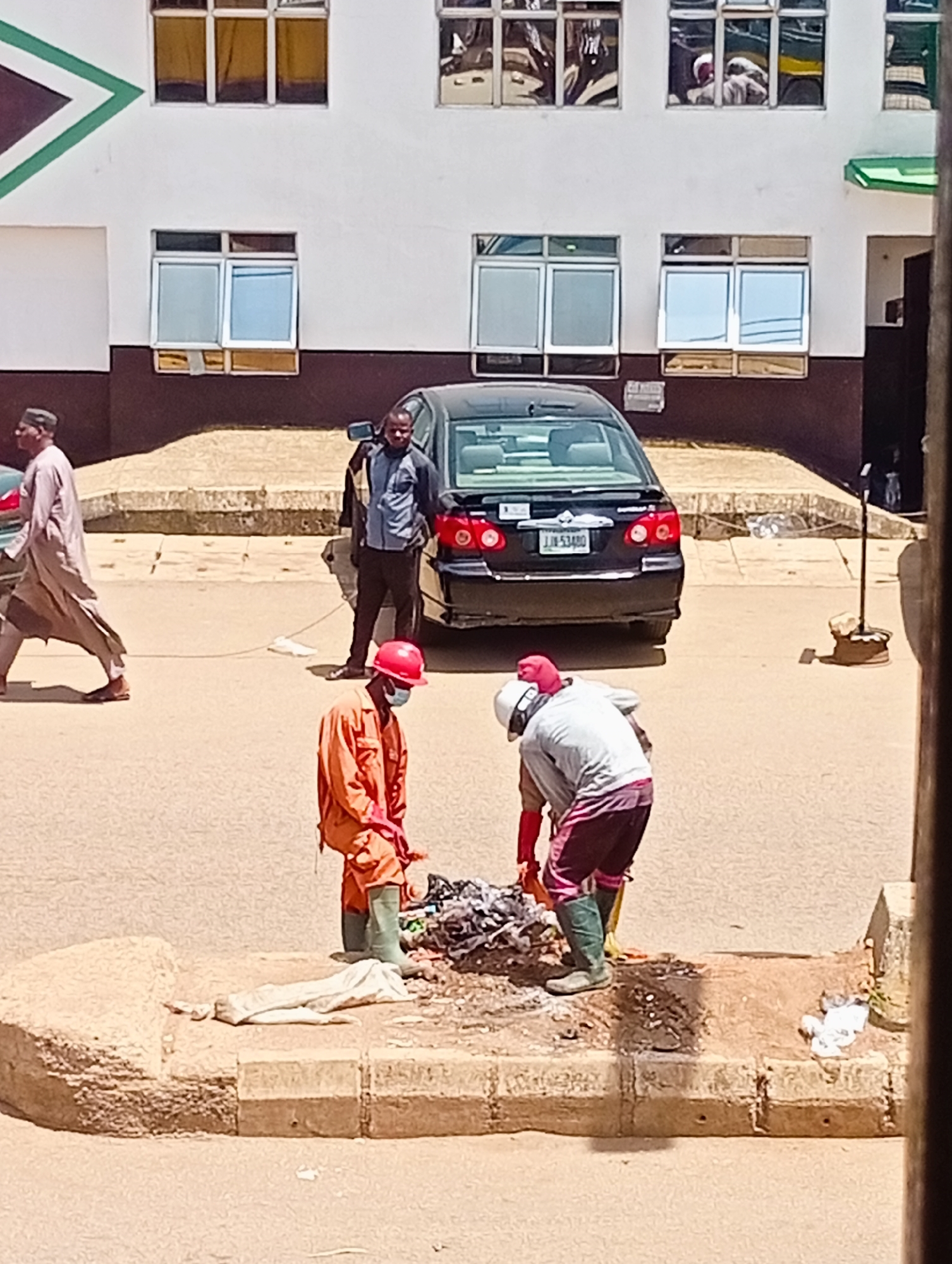 In Bukuru, Nigeria, two individuals wearing protective gear are tending to a pile of debris on a dusty roadside. The person on the left, dressed in an orange jumpsuit and a red helmet, is bent over, seemingly collecting or sifting through the waste. The person on the right, wearing a grey top, pink and white striped pants, and a white helmet, is also engaged with the debris. Adjacent to them, a light-colored cloth or garment lies on a concrete curb. In the background, a black sedan is parked, and a man in a blue shirt and dark pants stands beside it. Further back, a white building with multiple windows is visible. The ground is paved and dusty, characteristic of a street environment.
