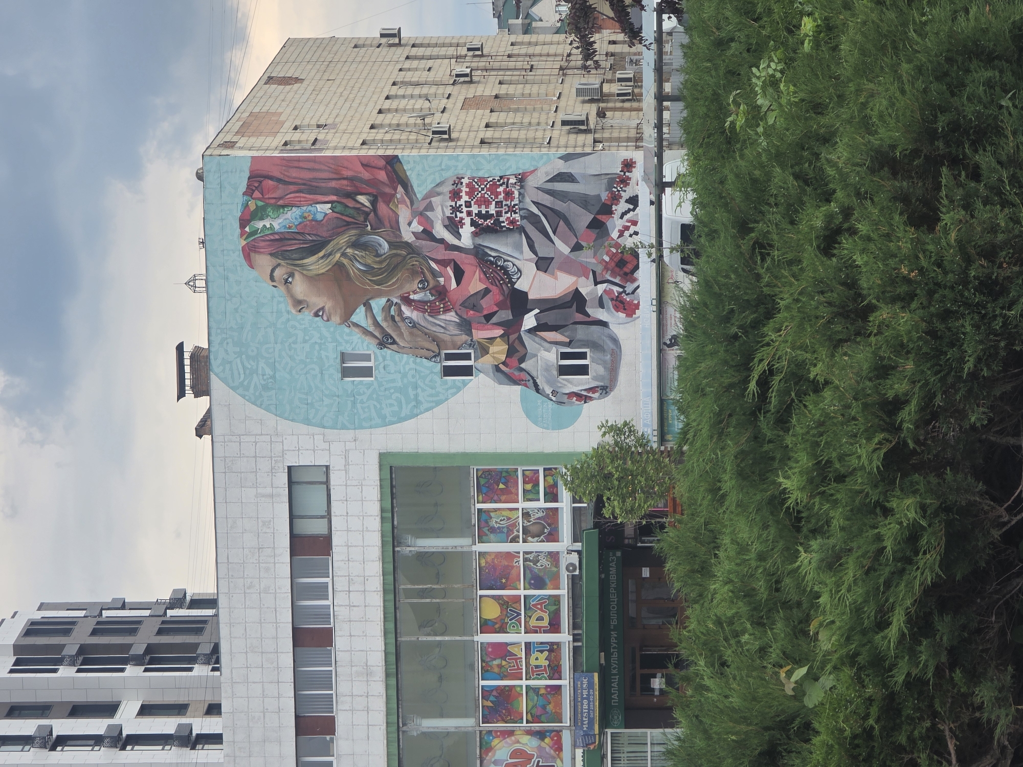 An outdoor, eye-level, full shot captures a vibrant mural on a multi-story building in Bila Tserkva, Ukraine, under an overcast sky. The mural, located on the left side of the building's facade, depicts a woman's upper body and head in profile, looking upwards and to the left. She wears a red headscarf with green and floral patterns, a prominent necklace, and rings on her fingers. Her attire features geometric patterns resembling traditional Ukrainian embroidery (vyshyvanka style) in shades of red, black, white, and gray, set against a light blue background with subtle lighter blue patterns.
The building itself, identified by a sign reading "ПАЛАЦ КУЛЬТУРИ "БІЛОЦЕРКІВМАЗ"" (PALACE OF CULTURE "BILOTSERKIVMAZ") above its main entrance, has a light gray, tiled facade on its upper sections. Below the mural, large ground-floor windows display colorful "HAPPY BIRTHDAY" decorations. A smaller sign to the left of the main entrance advertises "МУЗИЧНИЙ МАГАЗИН MAESTRO MUSIC" (MUSIC STORE MAESTRO MUSIC). The right side of the building, perpendicular to the mural wall, is constructed of light-colored bricks and features multiple windows and numerous external air conditioning units.
In the upper left background, a modern, multi-story building with a white and gray facade and balconies is partially visible. Power lines traverse the mid-ground horizontally. The immediate foreground is dominated by dense, dark green manicured bushes and shrubs, partially obscuring the base of the buildings. A small white air conditioning unit is visible above the "HAPPY BIRTHDAY" windows, and a white van is partially visible behind the bushes on the right.