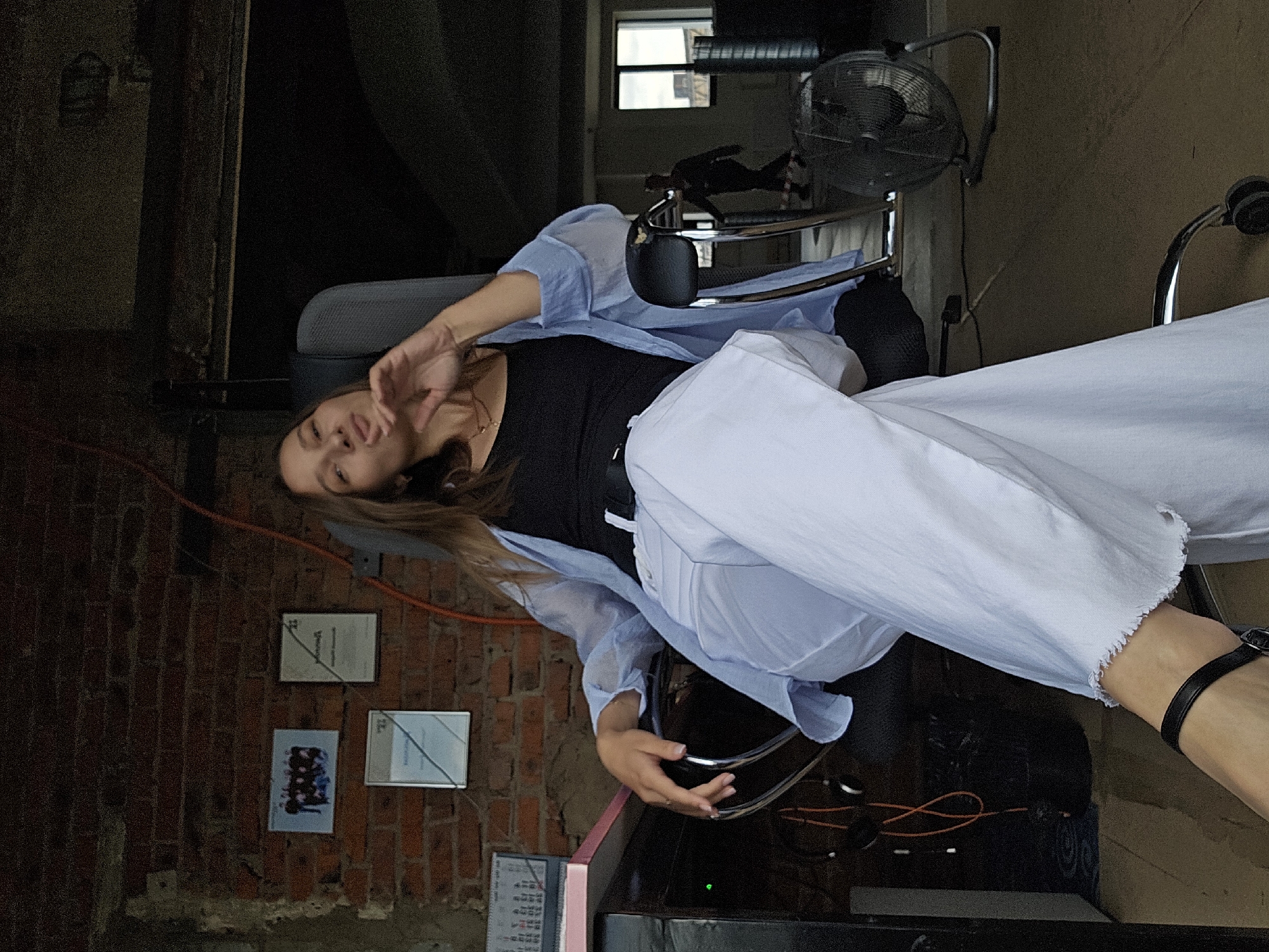 A young woman with long brown hair is sitting in a black office chair in a dimly lit room with exposed brick walls. She is wearing a light blue, loose-fitting blouse over a black tank top and white pants. Her left arm is resting on the armrest of the chair, and her right hand is raised to her face, touching her chin. She is looking directly at the camera with a playful expression.
In the background, there are framed pictures and documents on the brick wall. To the left, a desk with a pink surface and a calendar is visible. To the right, a person is walking in the distance, and a fan is positioned on the floor. The overall impression is a casual, candid shot taken indoors. There is no visible indication of the specific location within Chaikovsky, Russia.
