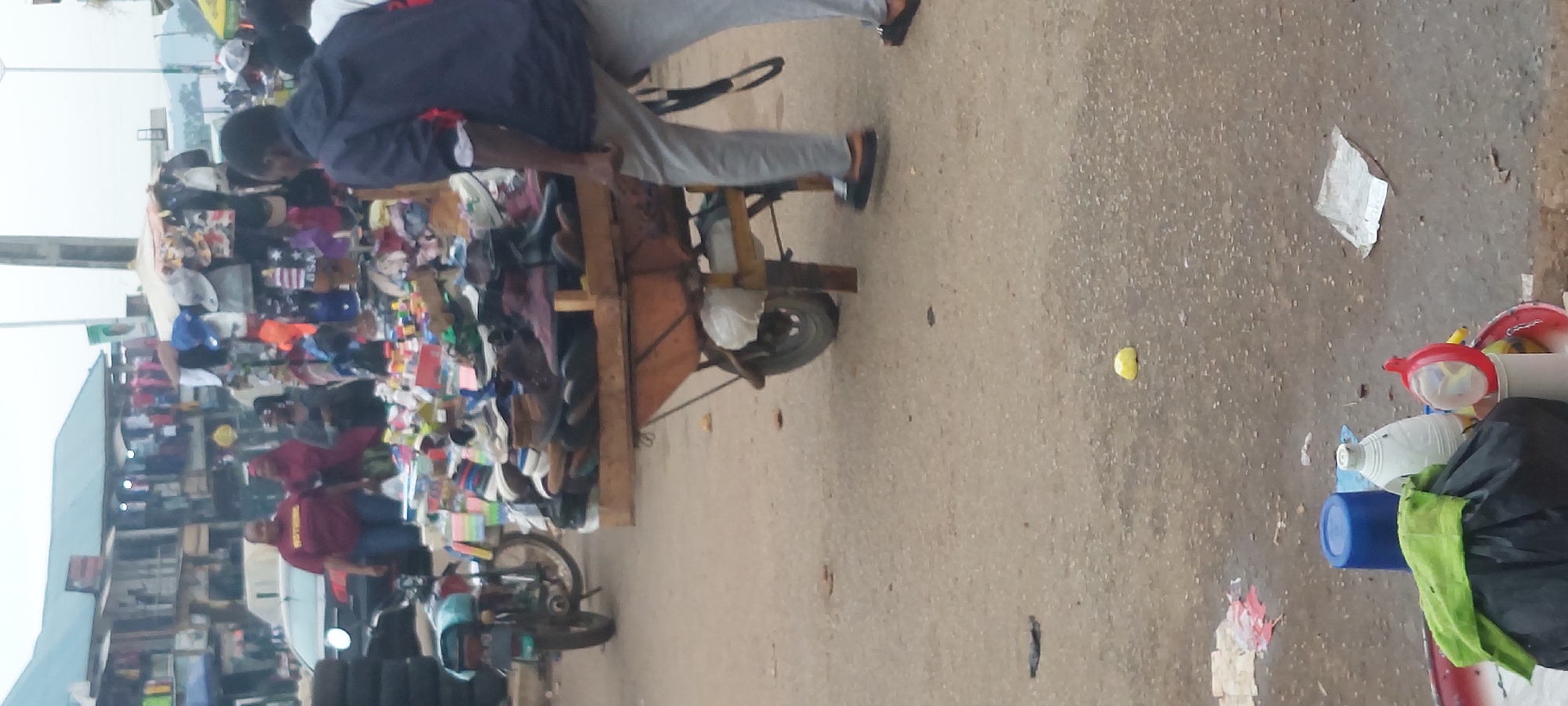 A street market scene unfolds in an outdoor urban environment. In the foreground, a man dressed in a dark blue hooded jacket and grey pants is pushing a wooden cart laden with various shoes. To the left, a motorcycle is parked on the asphalt ground, which is littered with debris, including a yellow object resembling a lemon, a crumpled silver package, and a discarded plastic bag. Further back, several individuals are visible. On the left, two women are standing near a car, one wearing a burgundy shirt with yellow text and the other a burgundy headscarf. To their right, another woman in a grey top is looking away. Behind them, stalls are set up with racks displaying an assortment of clothing items, including t-shirts, hats, and other apparel. The background shows buildings with multi-story structures and awnings, suggesting a busy commercial area. The sky is overcast, casting a diffused light over the scene.