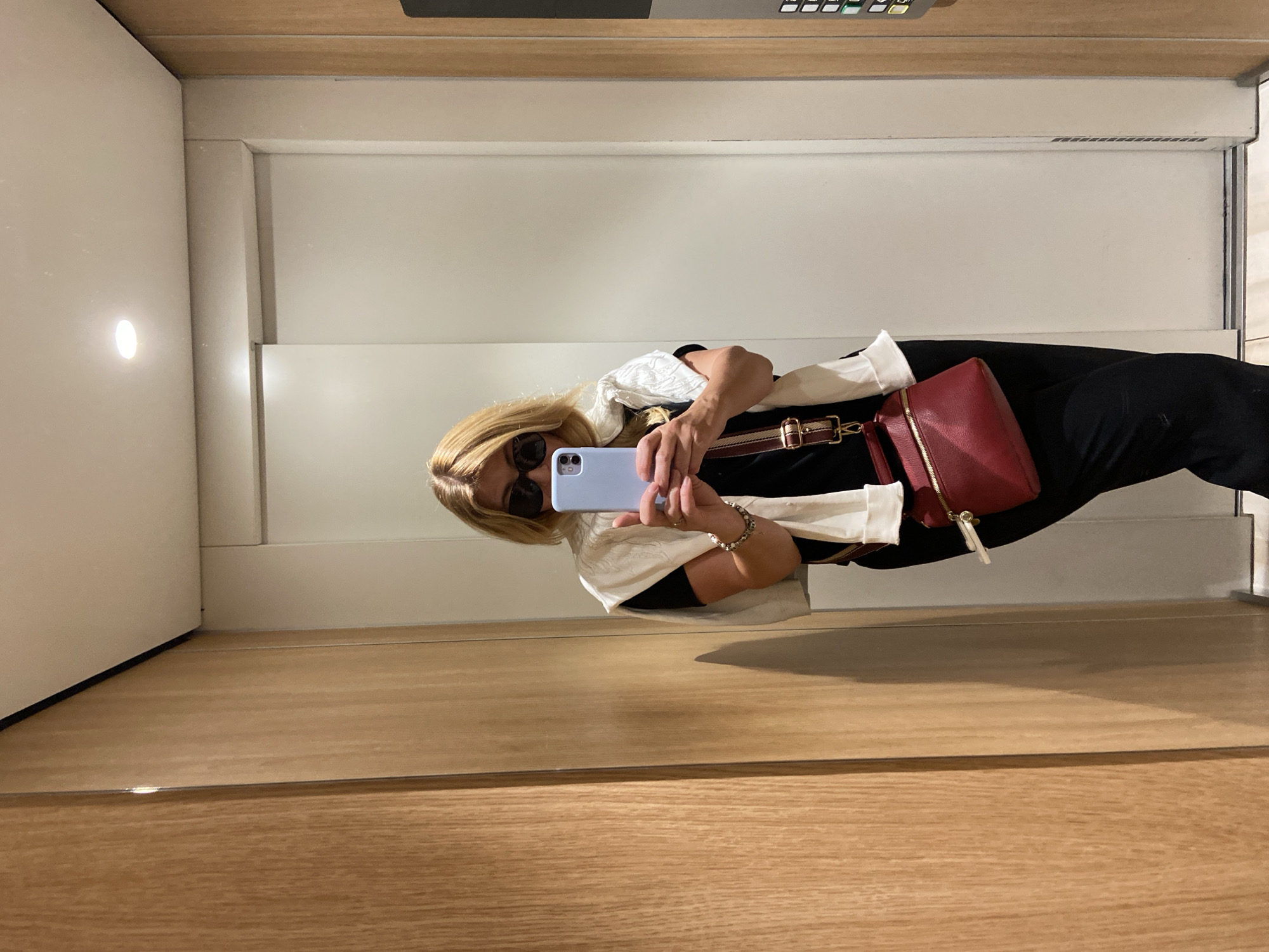 The image, rotated 90 degrees clockwise, captures a person taking a mirror selfie in what appears to be a contemporary indoor setting. The scene features light-colored walls and a wooden floor or lower wall panel, all reflected in a large mirror that the subject is facing.
The primary subject is an individual with blonde hair, wearing dark sunglasses, a black garment, and a white or cream-colored open jacket or shawl. They are holding a light blue smartphone with both hands to take the selfie, with their face partially obscured by the phone and sunglasses. A red cross-body bag with a distinctive striped strap (featuring red, white, and brown colors) hangs across their body. A multi-beaded or metallic bracelet is visible on their right wrist.
In the background, to the left of the subject's reflection, a small, circular wall-mounted light fixture casts a soft glow. Above the reflection, a panel with several illuminated buttons or indicators suggests a control panel, possibly found in an elevator or a modern interior space. The setting is well-lit, likely by artificial light sources. No discernible text is visible on any objects or surfaces within the frame. The overall atmosphere suggests a casual moment of self-documentation in a clean, functional environment.