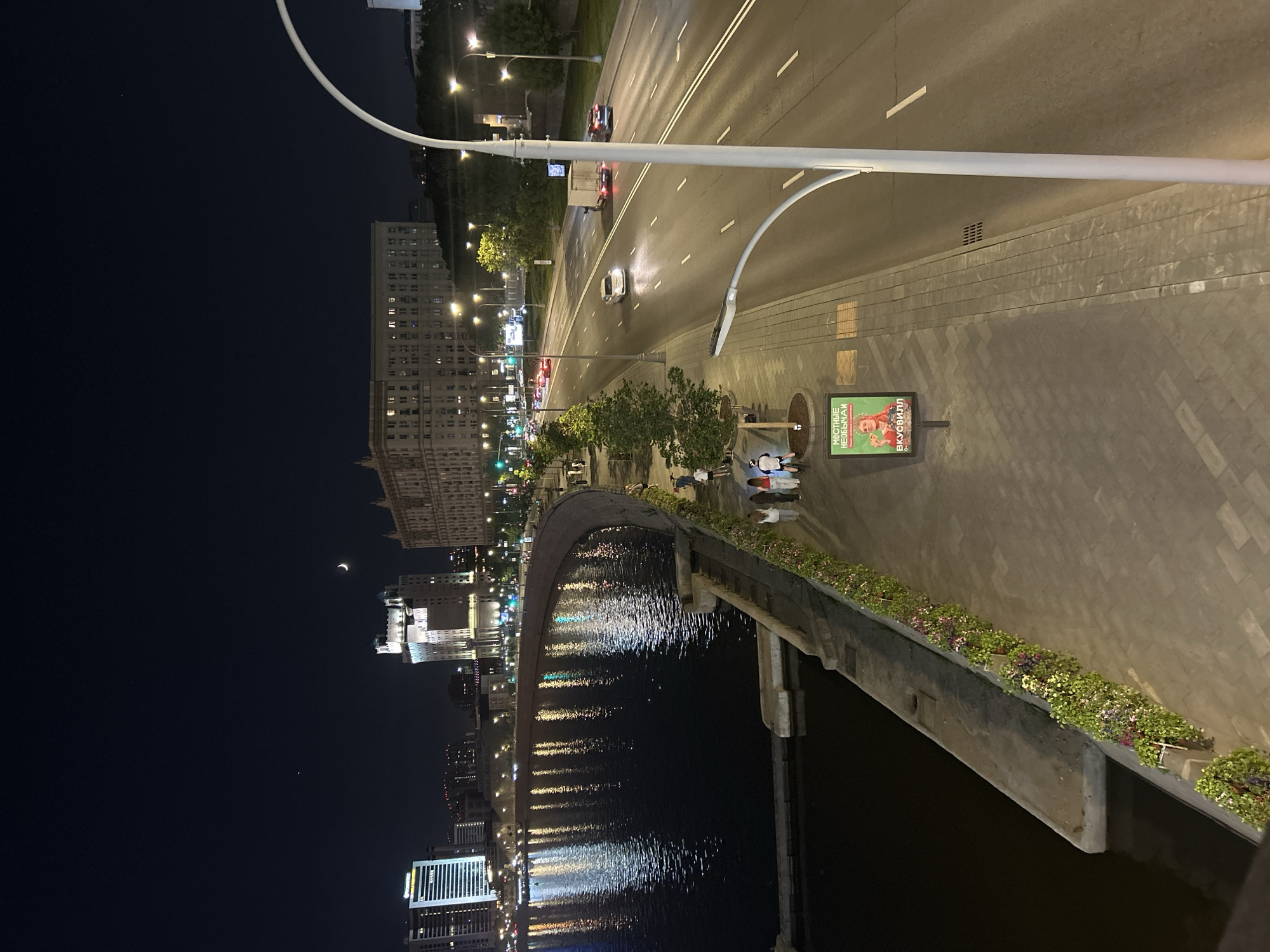Here is a description of the image:
The image depicts an urban landscape at night. In the foreground, a bridge spans a body of water, with reflections of city lights shimmering on the surface. A sidewalk runs alongside the road. People are strolling along the sidewalk. The sidewalk is edged with planters filled with flowers. A billboard stands near the sidewalk. Beyond the sidewalk is a road. The road contains vehicles driving on it. Buildings line the horizon, illuminated with lights. The sky is dark, with a crescent moon visible. The scene is lit by streetlights and building lights.