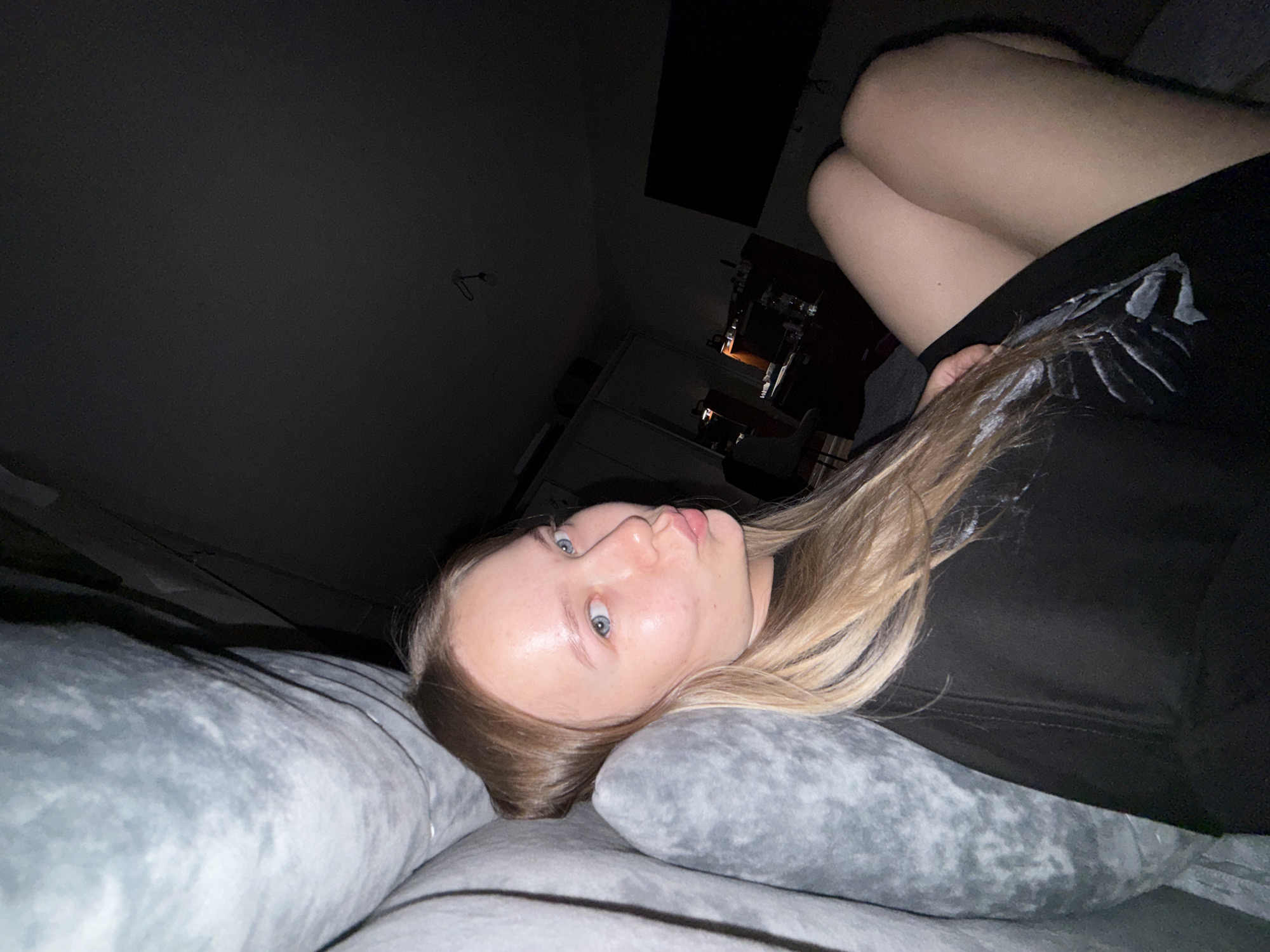 A young woman with light skin and blonde hair, lying on her side, is the central subject of this indoor photo. She has blue eyes and is looking directly at the camera with a neutral expression, her lips slightly parted. Her hair is spread out on a grey, plush pillow, suggesting she is in a bed or on a couch. She is wearing a dark-colored top, possibly black, which covers her torso and arms. Her bare legs are visible, bent at the knees and positioned towards the upper right of the frame, showing light-toned skin.
The lighting in the image is dim, indicating it might be evening or nighttime. The background is dark and somewhat indistinct, but hints of furniture and household items can be seen. On the left side of the background, there's a dark wall with what appears to be a light fixture or hook. Towards the center and right, there's a darker rectangular shape, possibly a window or a mounted object, and further down, the outlines of what could be a desk, chair, or other furniture items are vaguely discernible. There are warm, faint glows of light in the background, suggesting ambient lighting from lamps or electronic screens. The overall mood is quiet and relaxed. No visible text is present in the image. The location is an indoor setting, likely a bedroom or living room.