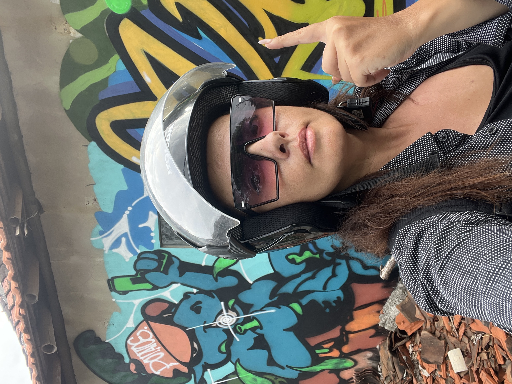 A medium shot captured from the chest up shows a woman taking a selfie in front of a graffiti-covered wall in Kuta Selatan, Indonesia. The woman, wearing a black helmet with a clear visor and pink-tinted sunglasses, points her right index finger upwards. She has long, brown hair and is wearing a black top under a black and white patterned button-up shirt. The background features a colorful graffiti mural with a blue cartoon character wearing an orange cap, and abstract yellow and green shapes. To the left of the woman, a pile of broken terracotta roof tiles and rubble is visible. The upper portion of the image shows a section of a tiled roof with some pipes.
