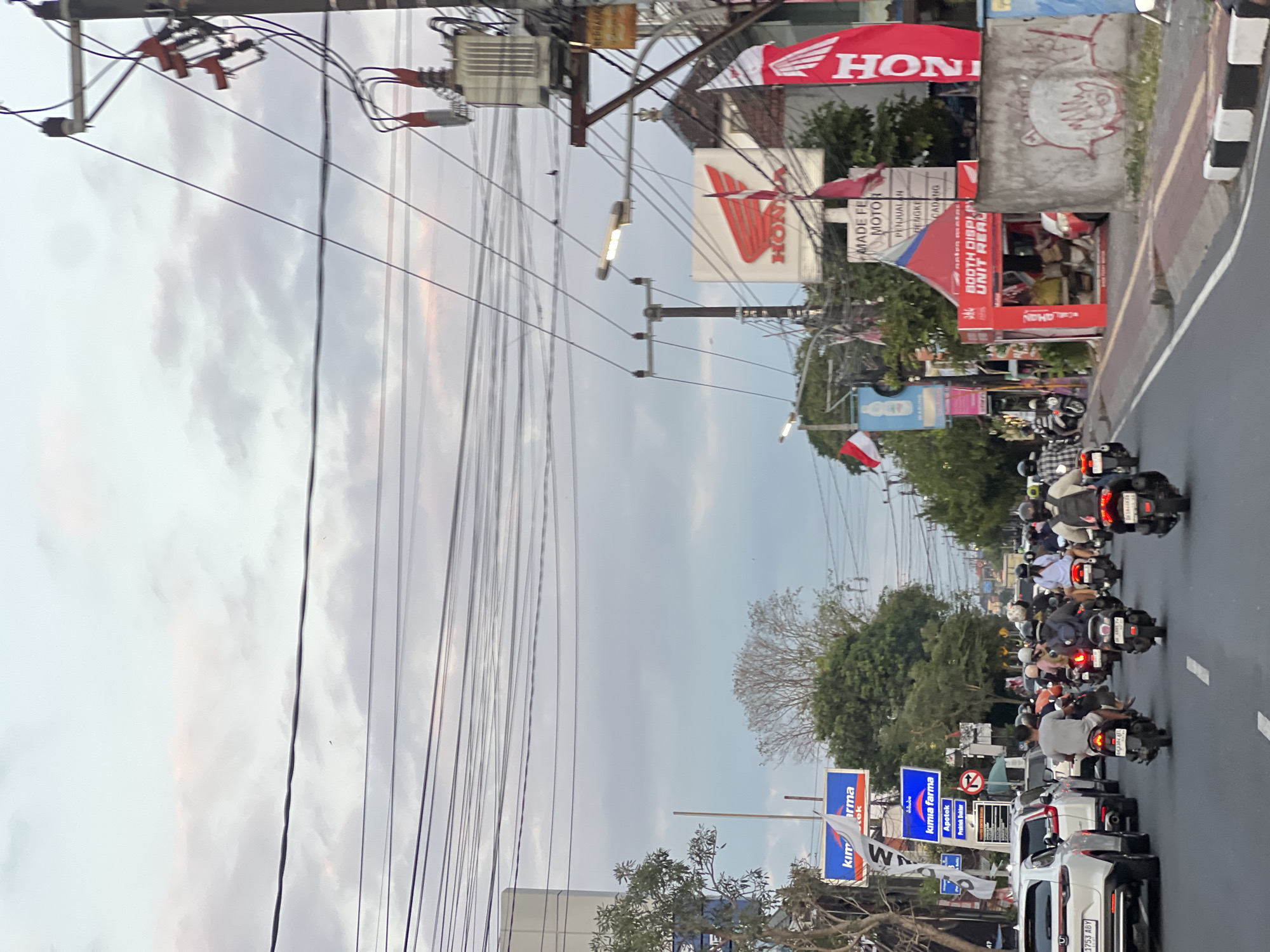 The image depicts a street scene in Denpasar, Indonesia, likely during the late afternoon or early evening, as indicated by the soft lighting and overcast sky. The street is busy with traffic, primarily motorcycles, with a few cars also visible. Many people are riding motorcycles, some wearing helmets. Several commercial establishments are visible along the street, including what appears to be a Honda dealership with prominent signage and flags. There are also advertisements for "kimia farma" (a pharmacy chain) and other businesses. Utility poles with a dense network of overhead wires dominate the upper portion of the image, contributing to the urban atmosphere. The sky is filled with clouds, with hints of pink and orange suggesting the setting sun. The overall impression is one of a typical bustling urban environment in Indonesia.