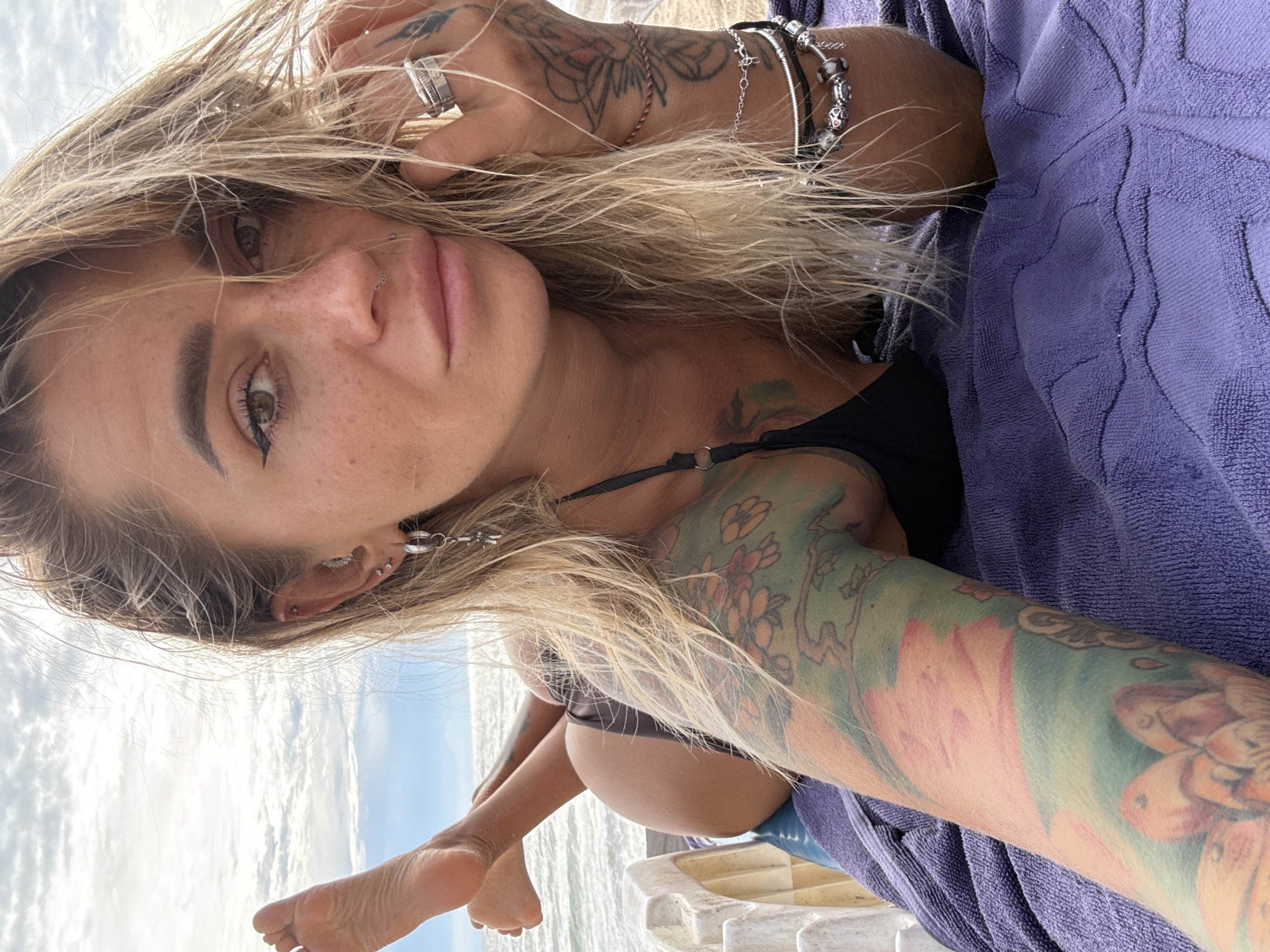 A woman with blonde hair and tattoos is lying on a purple towel on a beach. She is wearing a black bikini. Her feet are visible in the upper left corner of the image. The ocean and a cloudy sky are in the background. There is a white structure to the left of the woman.