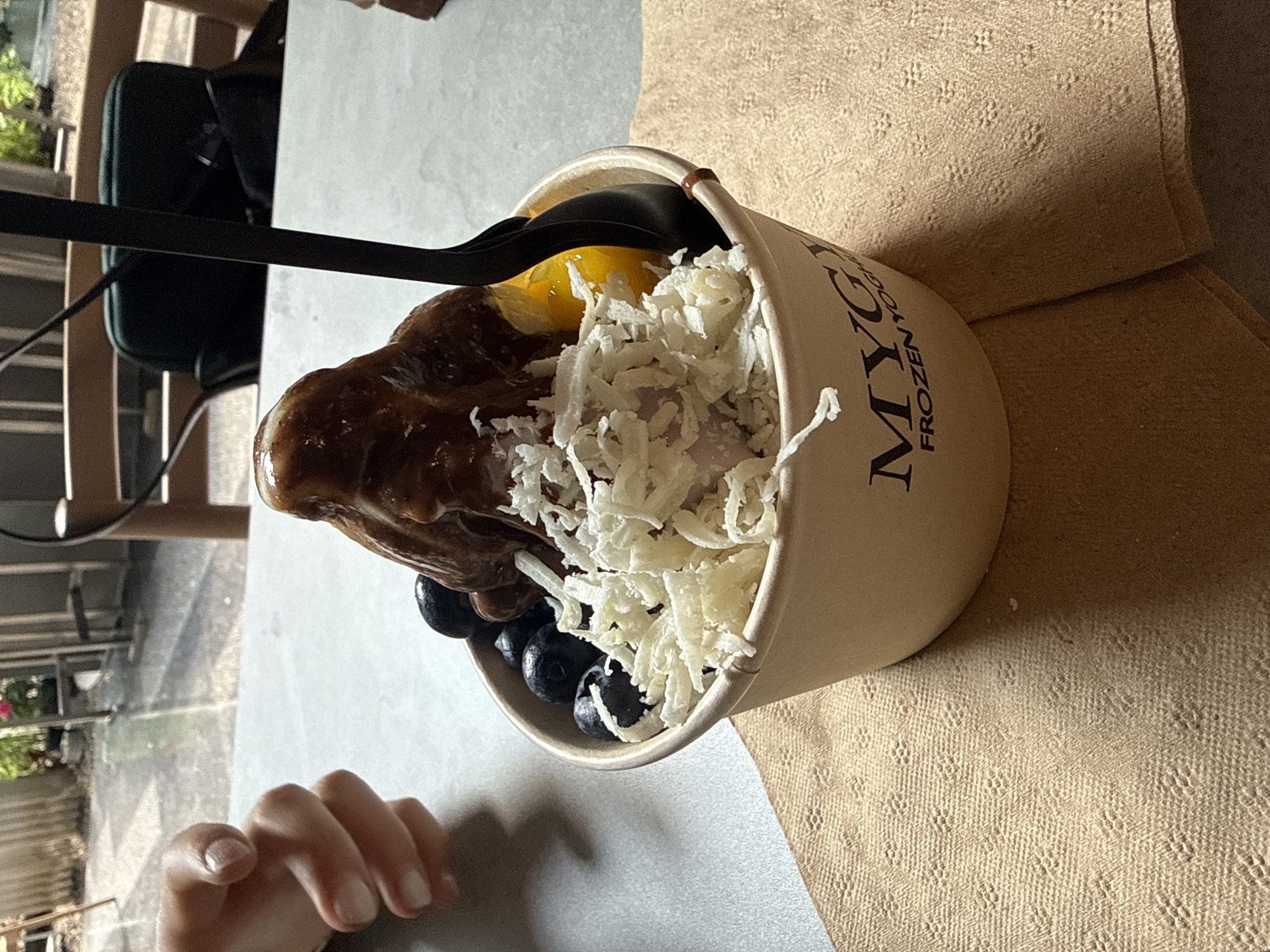 A close-up, angled shot depicts a cup of frozen yogurt from "MYGY FROZEN YOGURT," placed on a brown napkin or placemat. The cup contains a variety of toppings, including chocolate sauce, shredded coconut (or a similar white topping), blueberries, and what appears to be sliced mango. A black spoon rests inside the cup. A partial view of a hand with manicured nails is visible in the lower-left corner. The setting appears to be an outdoor seating area, potentially a patio, with glimpses of foliage and the exterior of a building visible in the background. The location is Mexico City, Mexico.