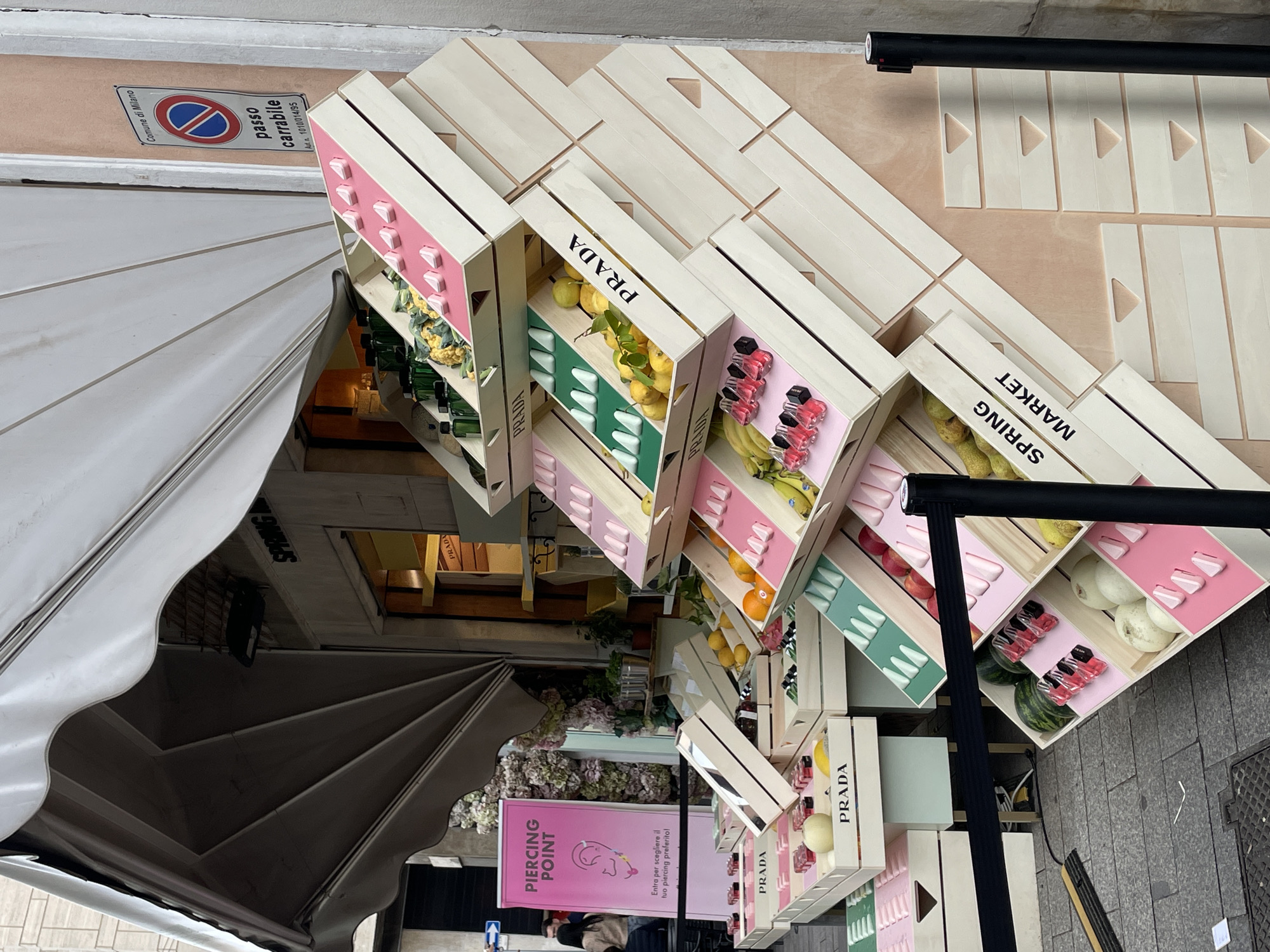 The image depicts a Prada "Spring Market" display, consisting of wooden crates containing various fruits, vegetables, and Prada beauty products, arranged in an eye-catching pattern outside a shop, presumably in Milan, Italy. The display is set up under a white awning, with a "Piercing Point" shop sign visible. A "Passo Carrabile" sign is also visible, indicating a designated driveway or access point in Milan. A couple of people are nearby. The overall scene suggests a promotional event or pop-up shop related to the Prada brand.