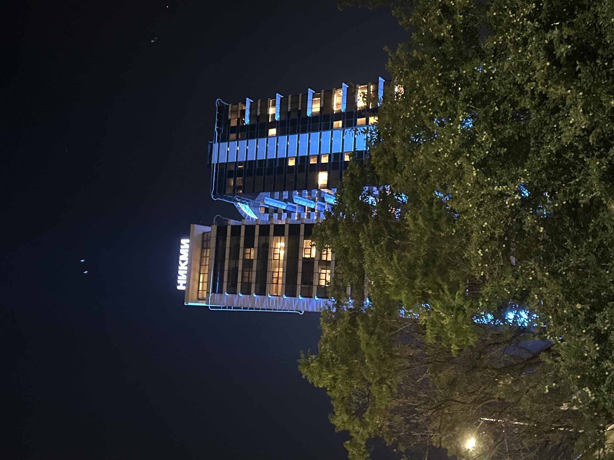 An exterior, low-angle, night shot shows two modern skyscrapers, one to the left and one to the right, behind a dense foreground of green trees. The skyscraper on the left is illuminated with a white sign that reads "НИКМИ" in white capital letters. Both skyscrapers are tall and rectangular with many windows. The skyscraper on the right is adorned with blue lights that streak down its facade, highlighting its verticality. The sky is dark blue and clear. In the distance, faint, small, white lights are visible in the sky, possibly distant aircraft or stars.